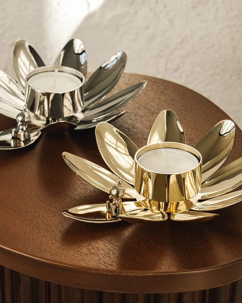 Zwei lotusförmige Teelichthalter, einer silber, einer gold, mit kleinen Buddha-Figuren, auf einem Holztisch.