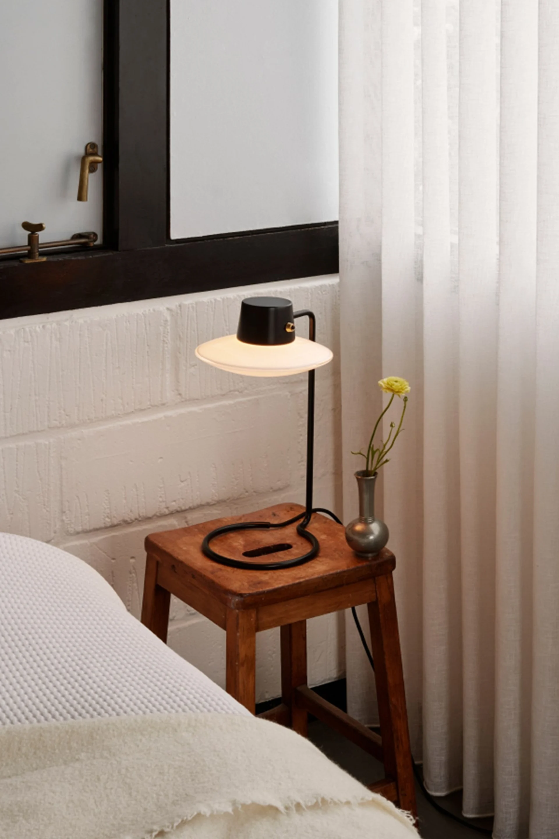 Schwarze Tischlampe mit breitem Schirm auf rustikalem Holzschemel, Vase mit gelber Blume, neben Bett und Vorhängen.