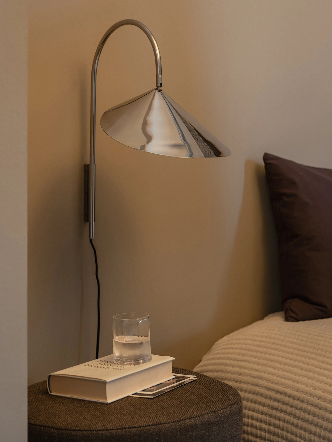 Eine moderne silberne Wandlampe mit konischem Schirm über einem Buch und einem Glas Wasser auf einem Nachttisch.