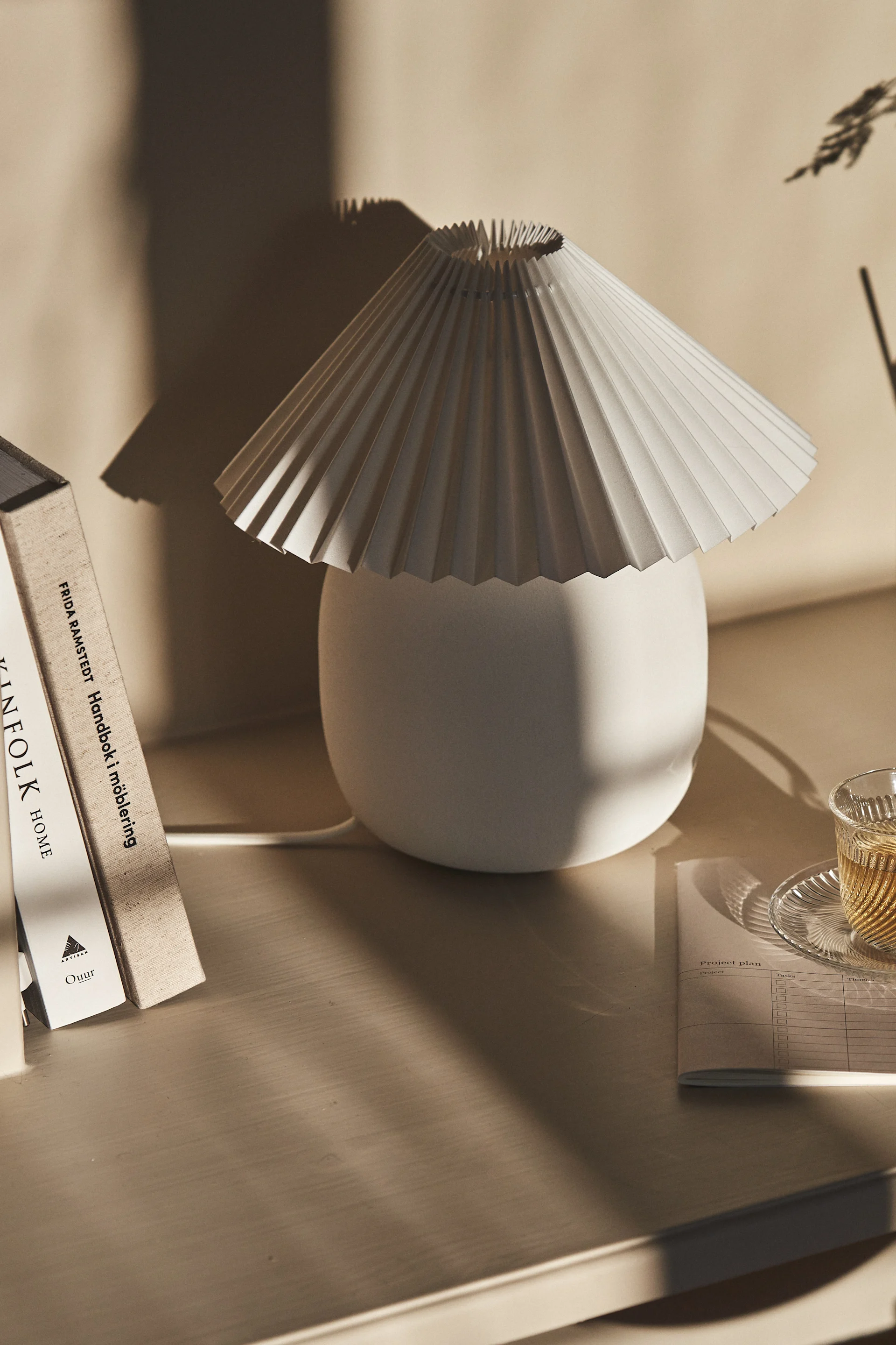 Eine plissierte weiße Tischlampe neben Büchern und einer Tasse Tee auf einem hellen Holzschreibtisch, mit weichen Schatten.