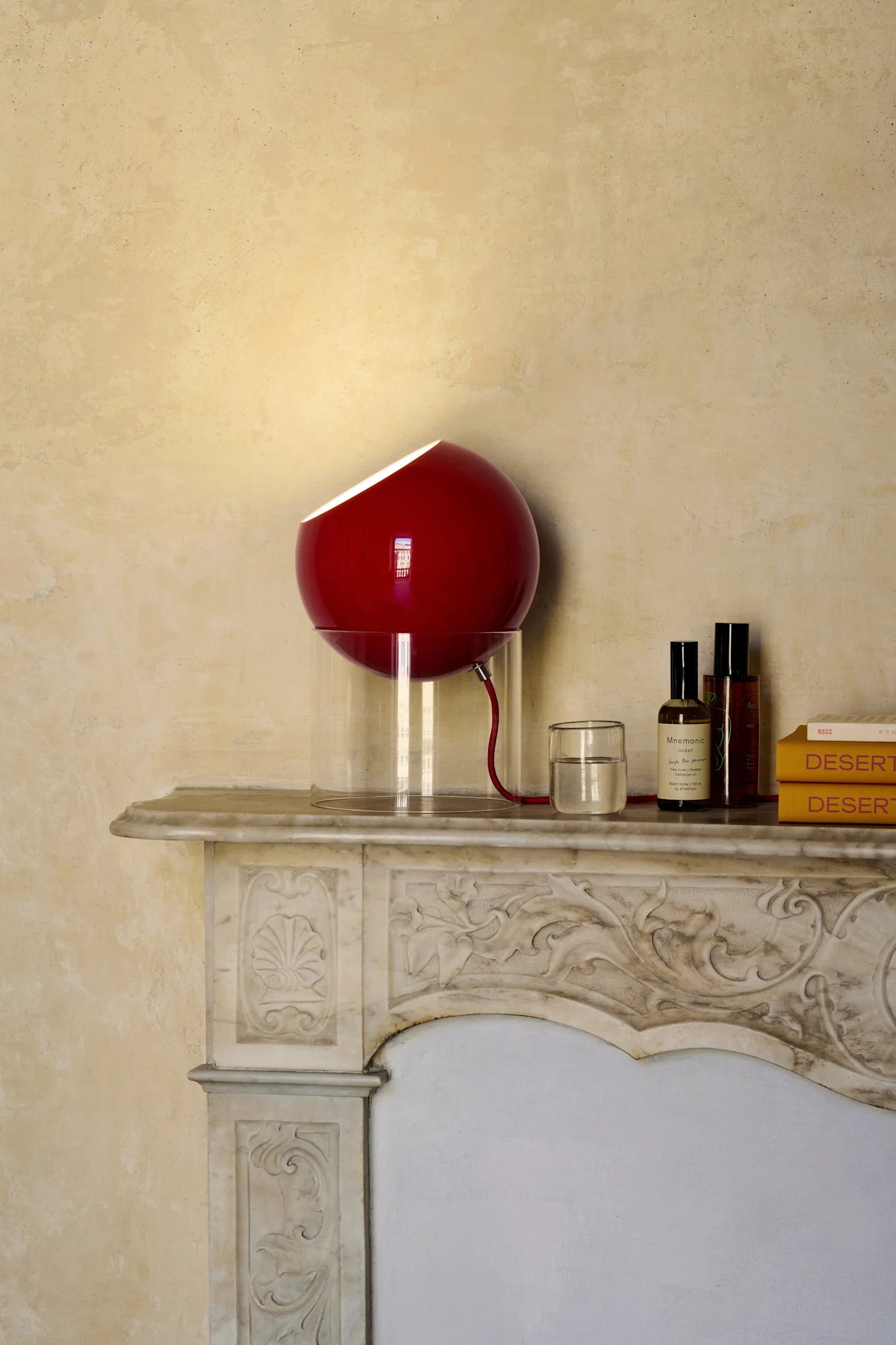 Rote kugelförmige Lampe mit durchscheinendem Fuß auf einem verzierten Marmorkamin, daneben ein Glas Wasser, zwei Flaschen und zwei gelbe Bücher.