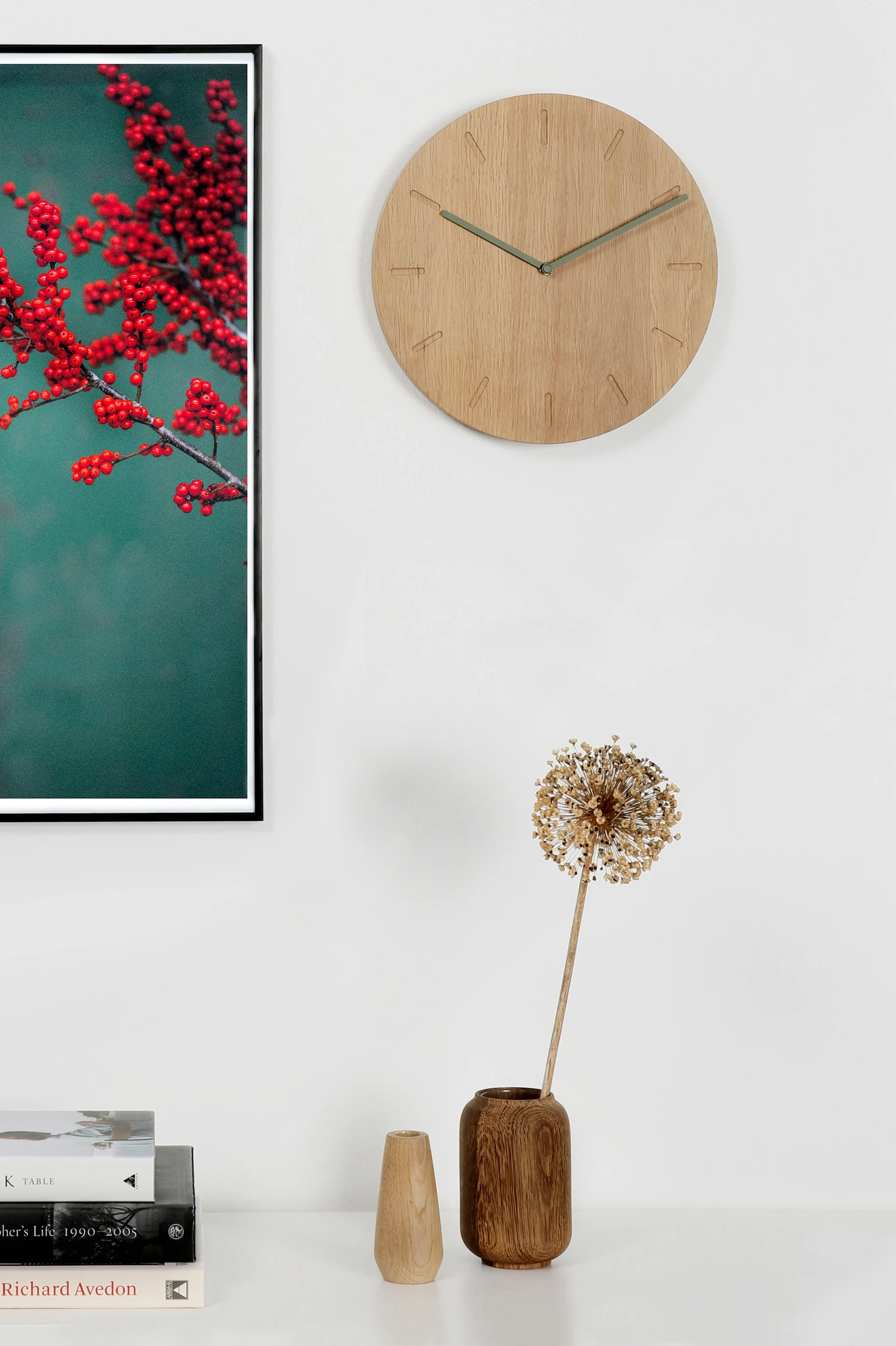 Ein gerahmter Druck mit roten Beeren auf türkisfarbenem Hintergrund hängt über einem Regal mit Büchern, einer Holzuhr und Trockenblumen in einer Vase.