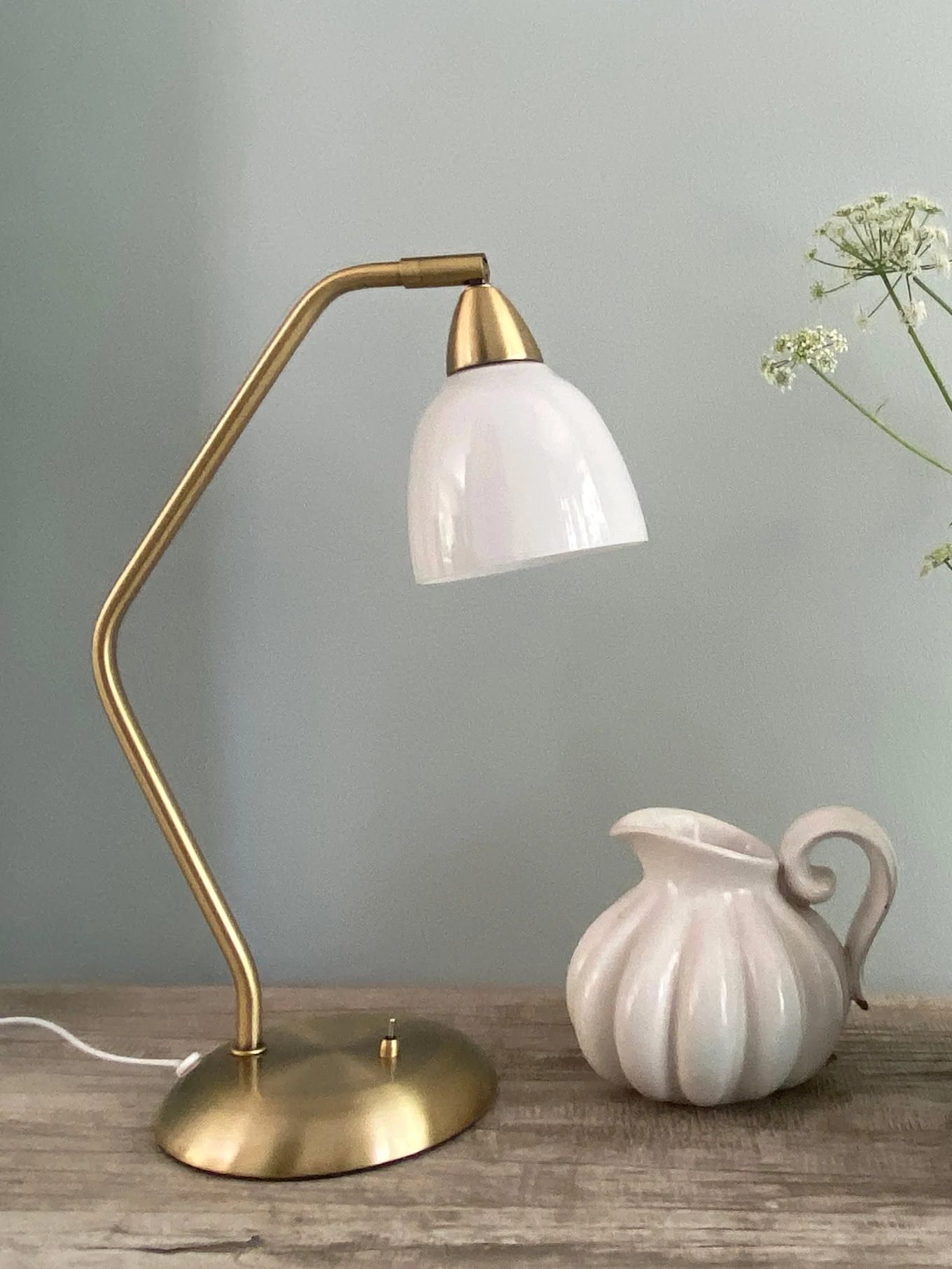Messingfarbene Tischlampe mit weißem Glasschirm und weißer geriffelter Keramikkaraffe auf Holzoberfläche.