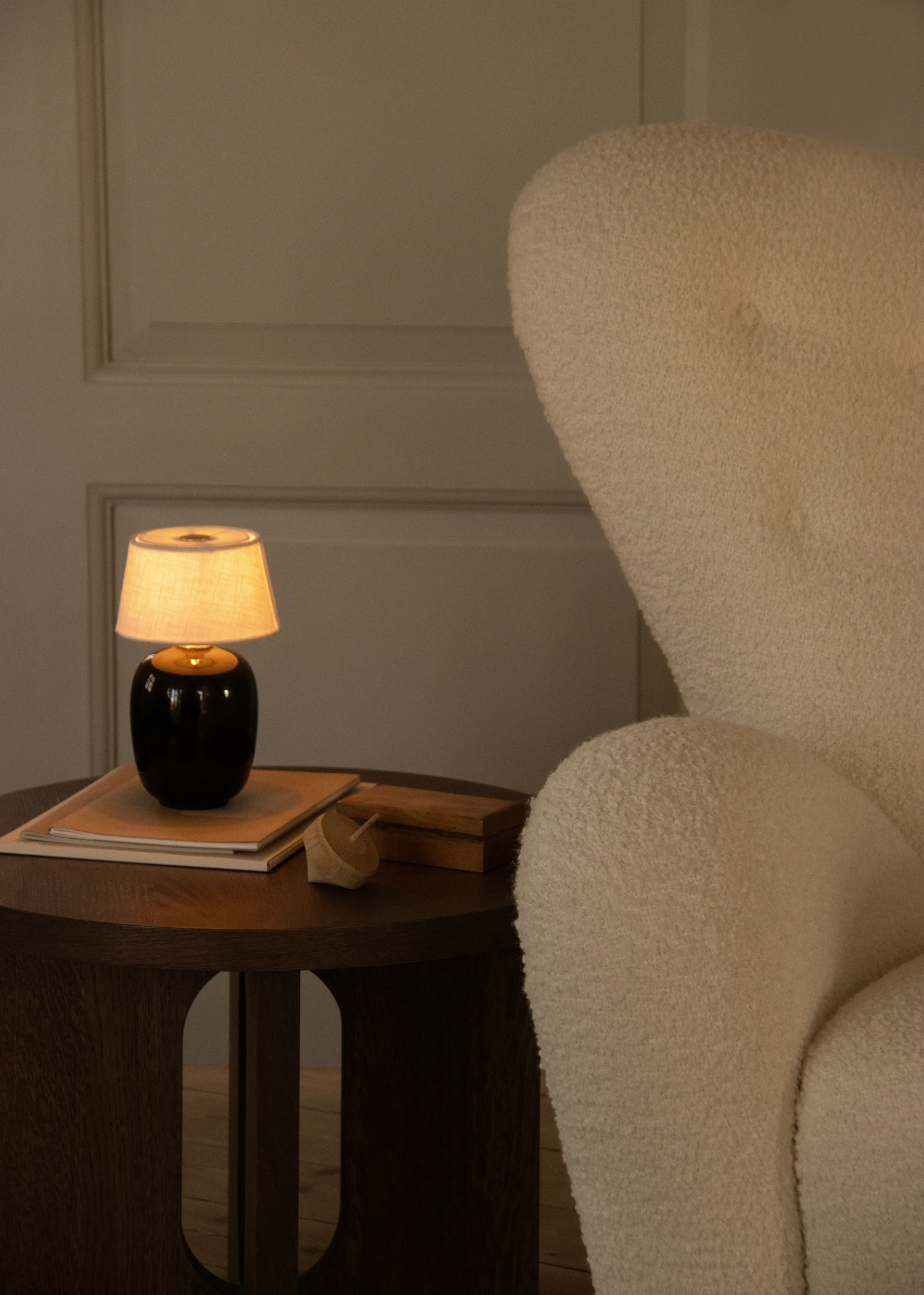 Warm beleuchtete gemütliche Ecke mit cremefarbenem Textursessel und Holztisch, darauf leuchtende Lampe, Bücher und Holzspielzeug.