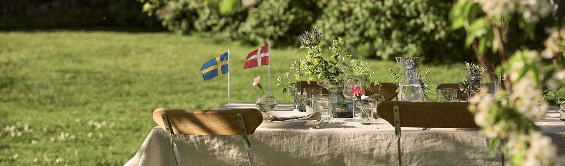 Gartentisch mit schwedischen und dänischen Flaggen, Wildblumen und Gläsern in einem sonnigen Garten.