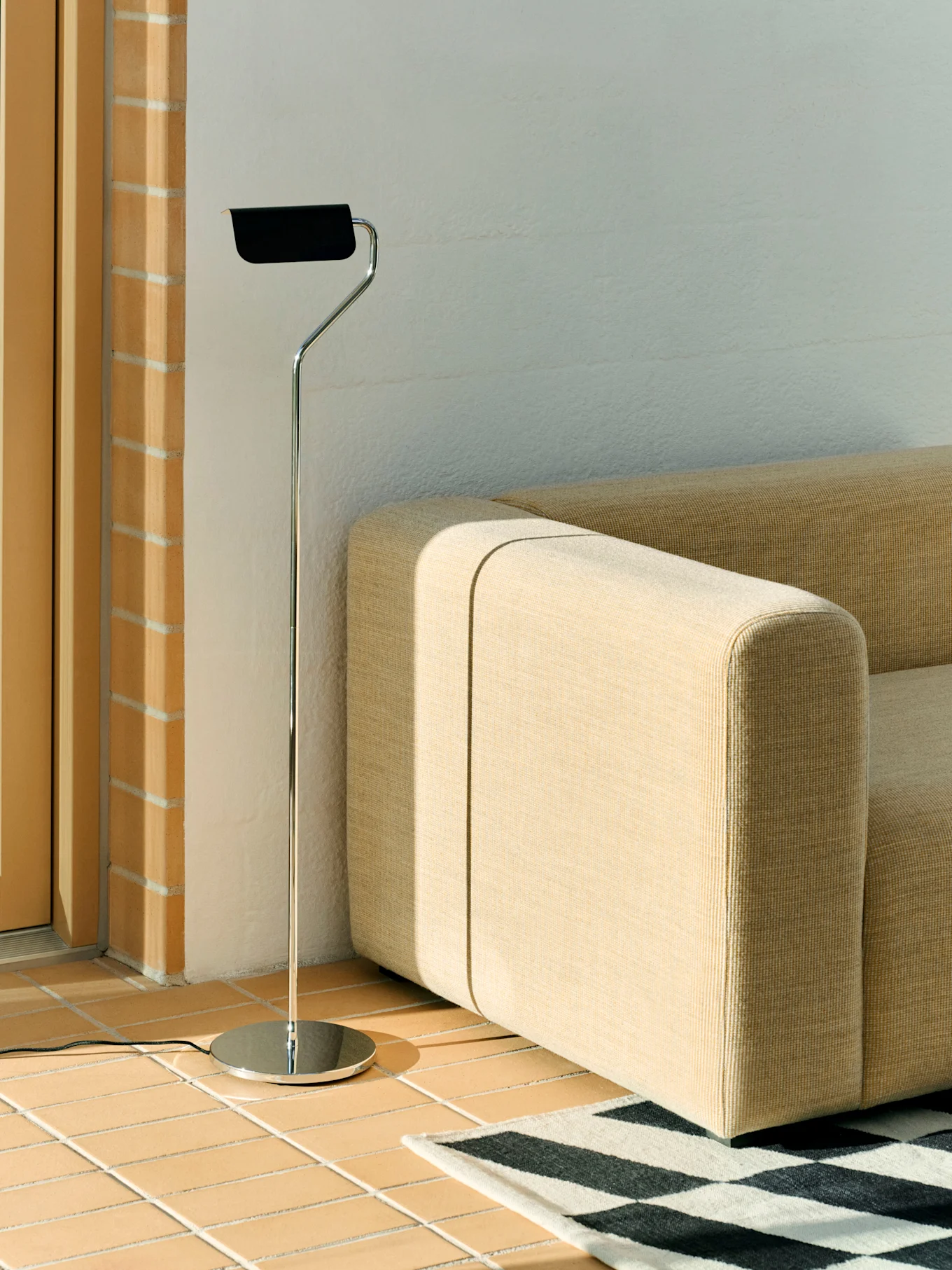 Eine Chrom-Stehlampe mit schwarzem Schirm steht neben einem beigen Modulsofa und einem schwarz-weiß gemusterten Teppich auf einem Fliesenboden.