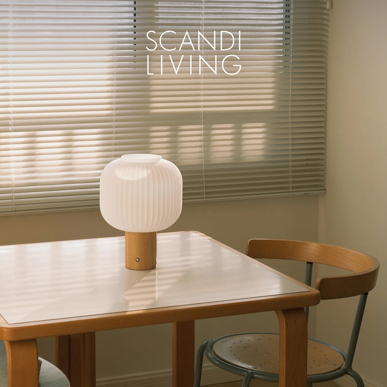 Weiße gerippte Tischlampe mit Holzfuß auf einem weiß gedeckten Tisch und einem Stuhl, vor Fensterjalousien. Text: SCANDI LIVING.