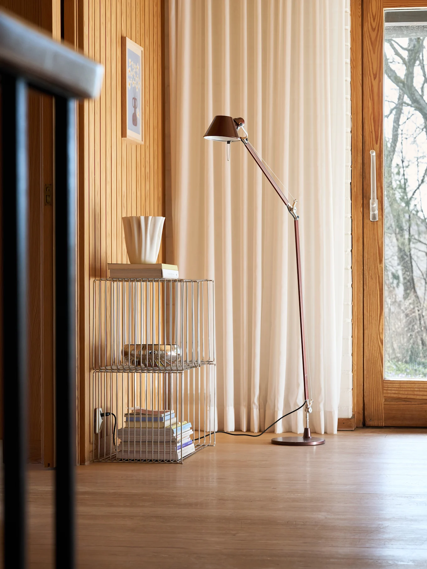 Ein gemütlicher Raum mit Holzwänden und -boden, einer roten Stehlampe, einem Chromregal mit Büchern und einer geriffelten weißen Vase sowie einem Fenster mit hellen Vorhängen.