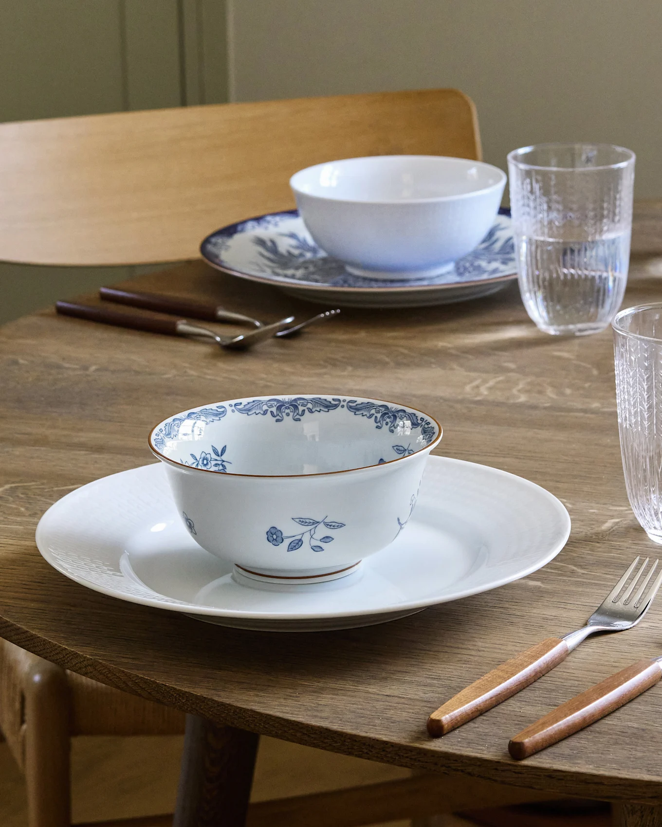 Esstisch für zwei Personen gedeckt mit weiß-blau gemusterten Schalen auf weißen Tellern, Besteck mit Holzgriffen und Wassergläsern.