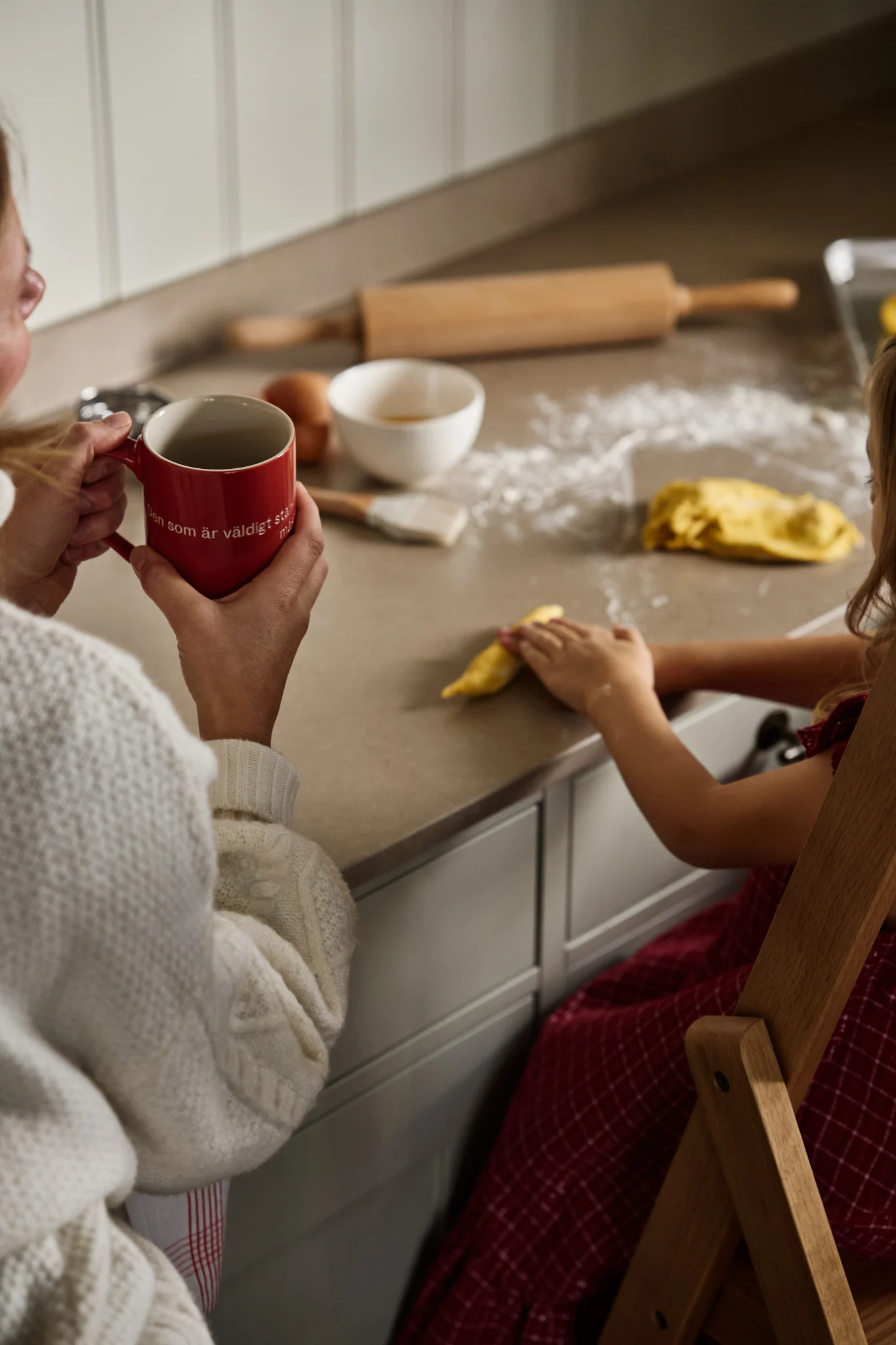 Eine Frau sitzt mit einer Astrid Lindgren Tasse an einer Küchentheke, davor ein Mädchen, das Teig rollt.