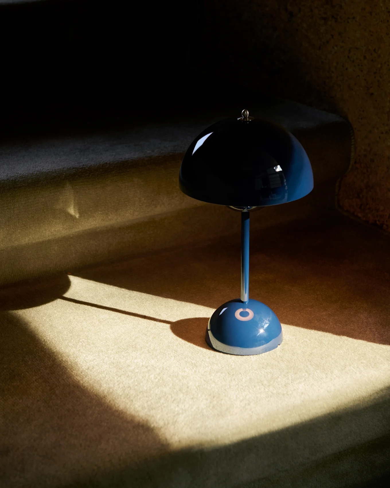 Eine blaue, pilzförmige Tischlampe mit reflektierendem Schirm steht auf einem Teppichboden, teilweise von Sonnenlicht beleuchtet.