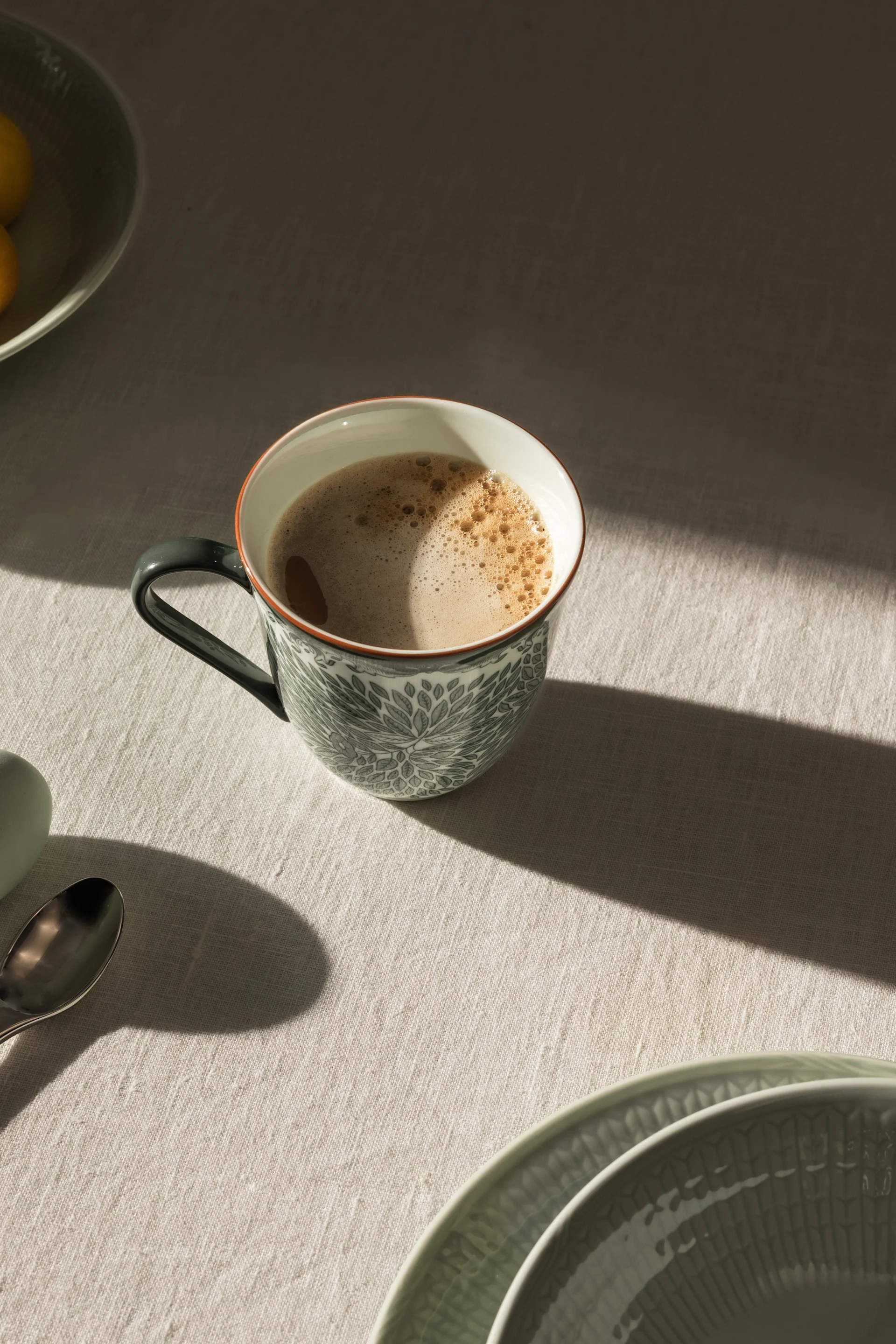 Eine gemusterte Keramiktasse mit schaumigem Kaffee auf einem hellen Tischtuch. Sonnenlicht wirft Schatten, daneben sind Zitronen, Teller und ein Löffel teilweise sichtbar.