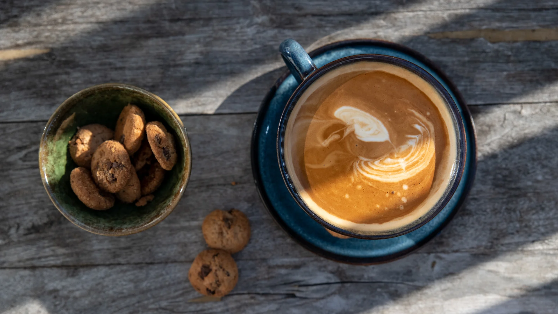 Aufnahme von oben: blaue Keramiktasse Kaffee mit Latte Art neben grüner Keramikschale mit Mini-Schokoladenkeksen auf Holztisch.