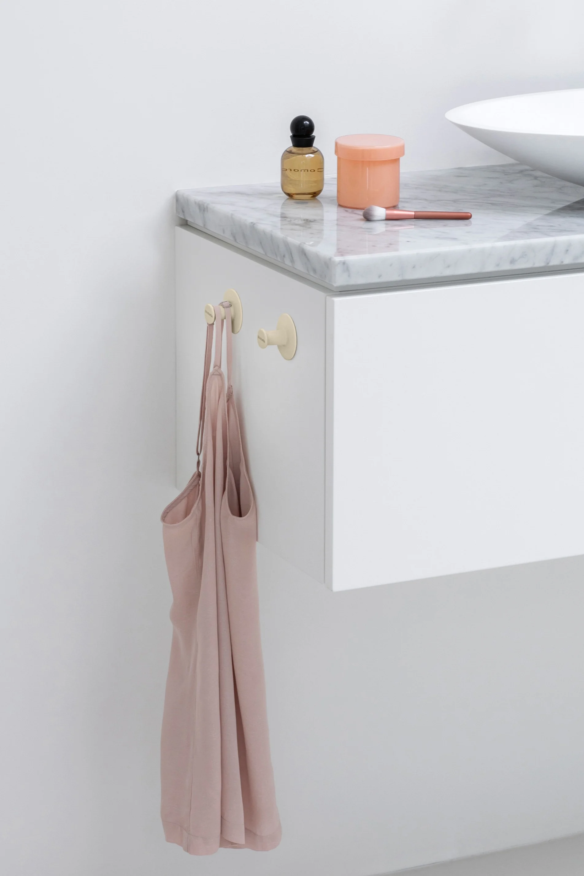 Ein hellrosa Camisole hängt an zwei runden hellgelben Haken an der Seite eines weißen Waschtischs mit Marmorplatte.