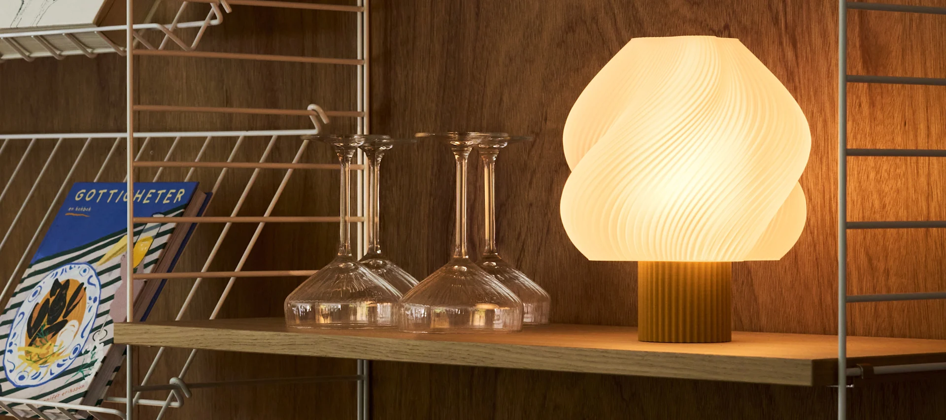 Eine beleuchtete, gerippte ovale Lampe und verschiedene klare Gläser stehen auf einem hölzernen Servierwagen vor einer warmen, strukturierten Wand.