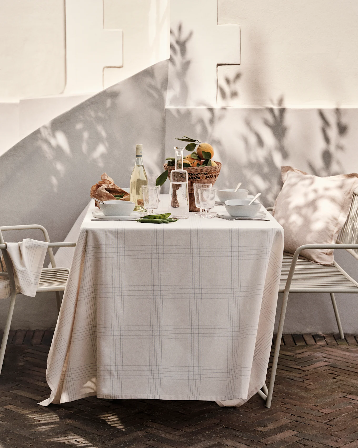 Draußen gedeckter Tisch mit karierter Tischdecke, Wein, Wasser, Schalen, Orangenkorb und Erbsen auf sonniger Terrasse.