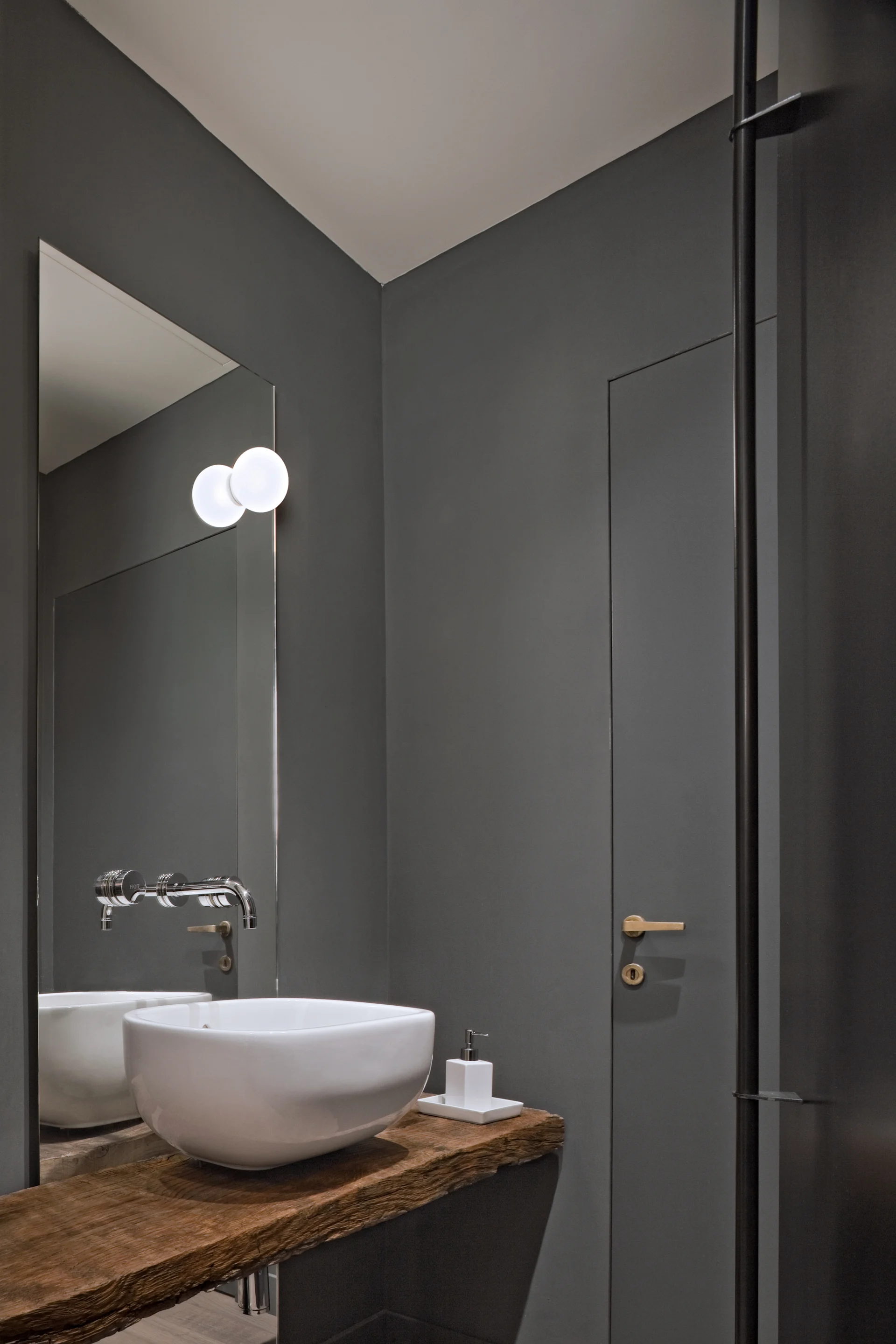 Modernes Badezimmer mit dunkelgrauen Wänden, einem weißen Aufsatzwaschbecken auf einer rustikalen Holzplatte, einem Spiegel und einem goldenen Türgriff.