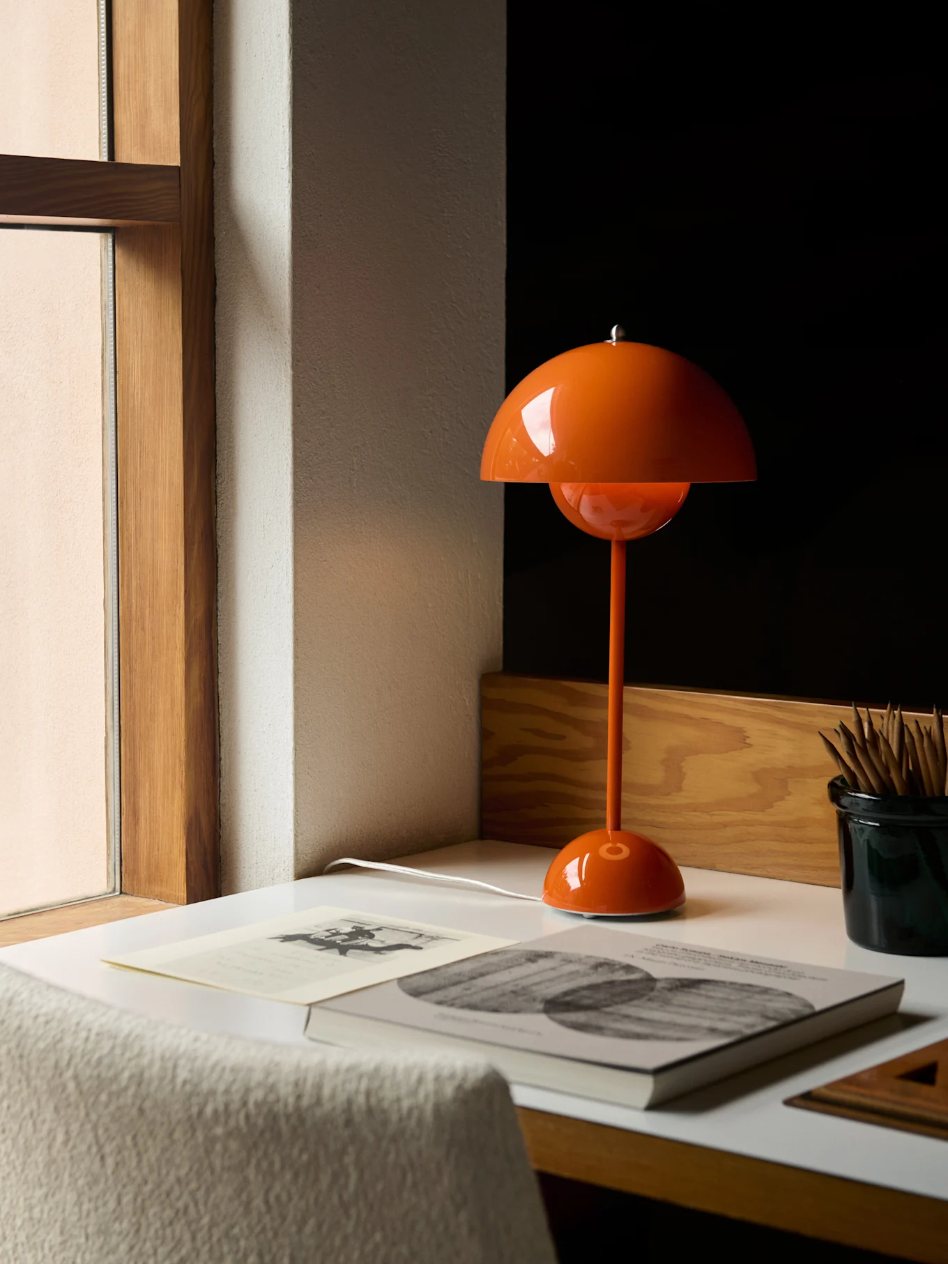 Eine orangefarbene, pilzförmige Tischlampe auf einem weißen Schreibtisch neben einem Fenster, mit Büchern und Bleistiften.