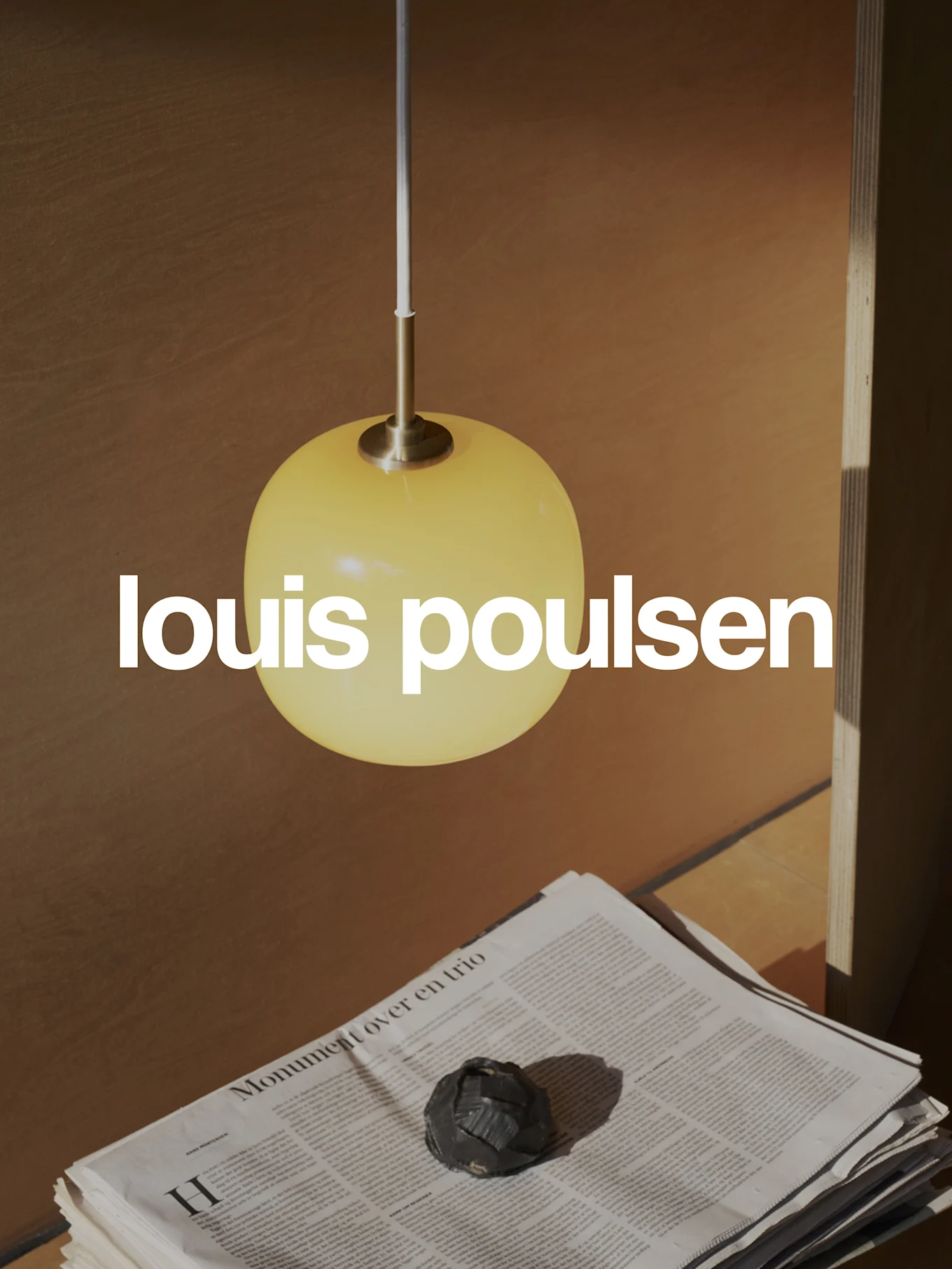 Louis Poulsen Logo über einer warmen Szene mit einer gelben kugelförmigen Pendelleuchte, gestapelten Zeitungen und einem dunklen Dekoobjekt.