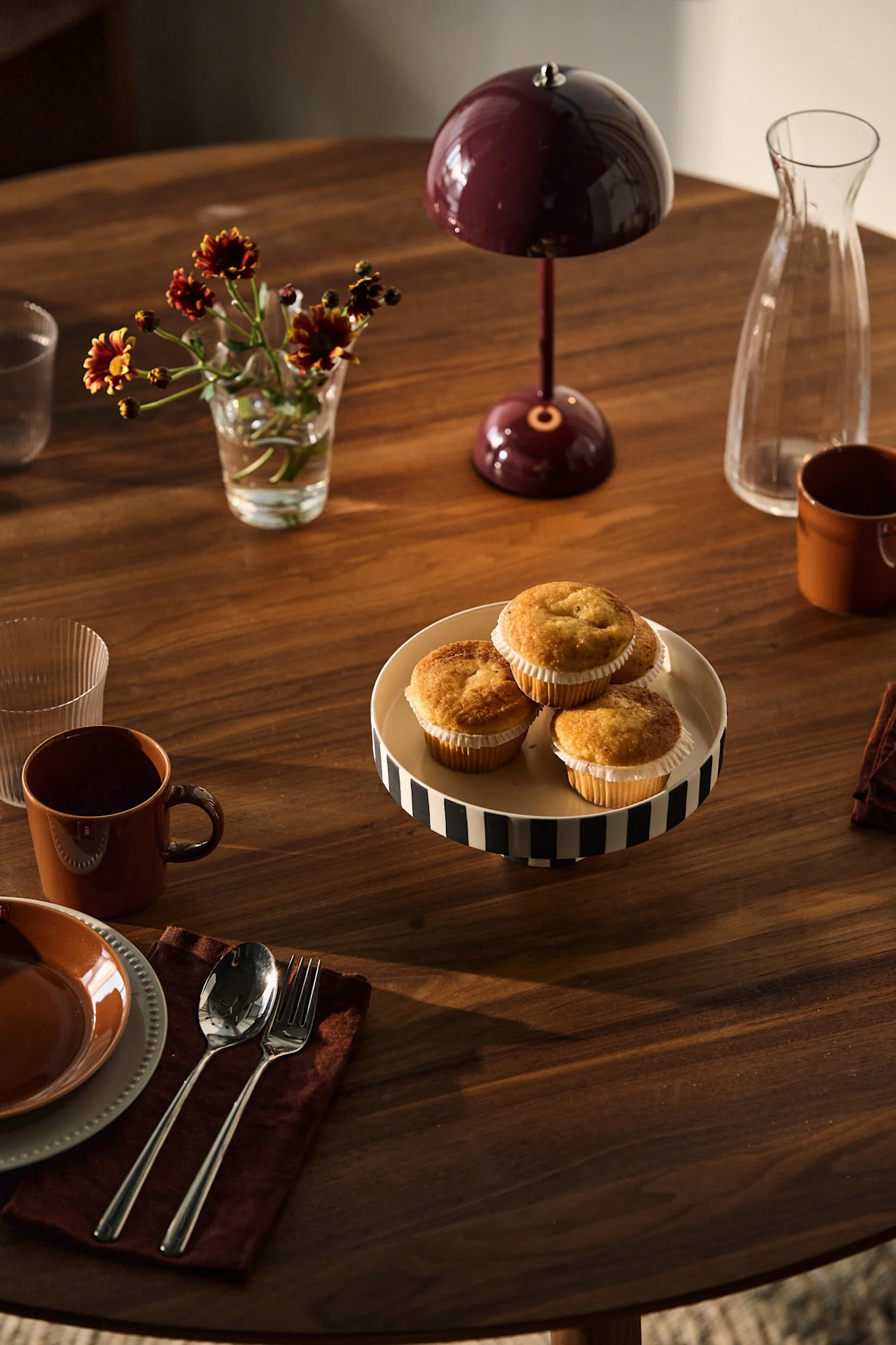 Ein Holztisch gedeckt mit Muffins auf einem gestreiften Kuchenständer, Blumen, der Akku-Tischleuchte Flowerpot von &tradition, Karaffe, Tassen und einem Gedeck.