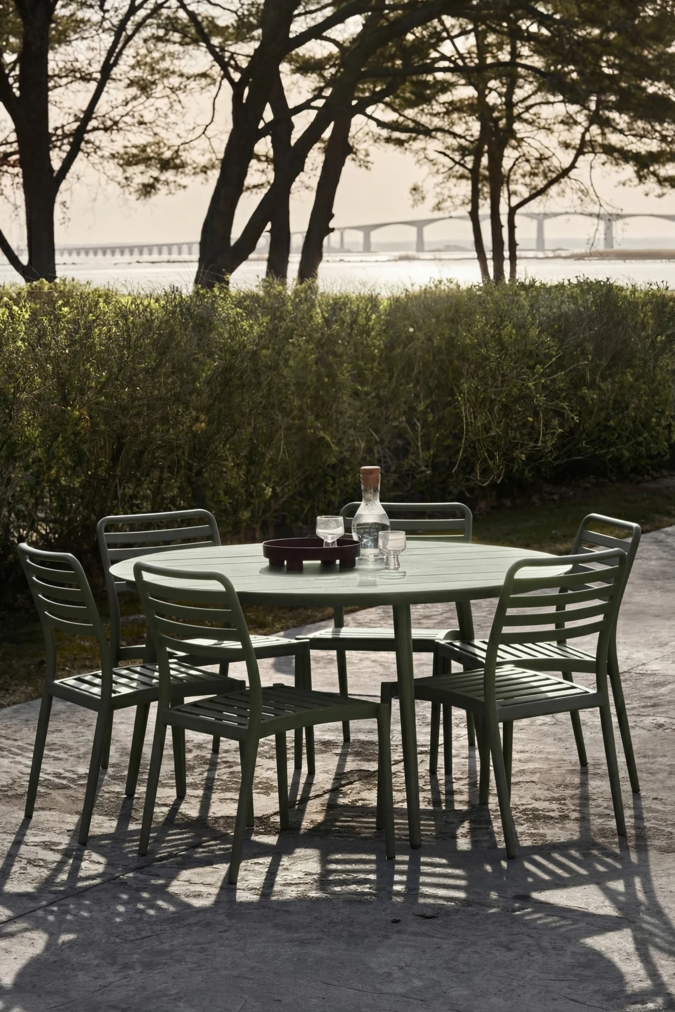 Grüne Outdoor-Essgruppe mit Tisch, fünf Stühlen, Wasserflasche und Gläsern auf der Terrasse. Brücke über Wasser.