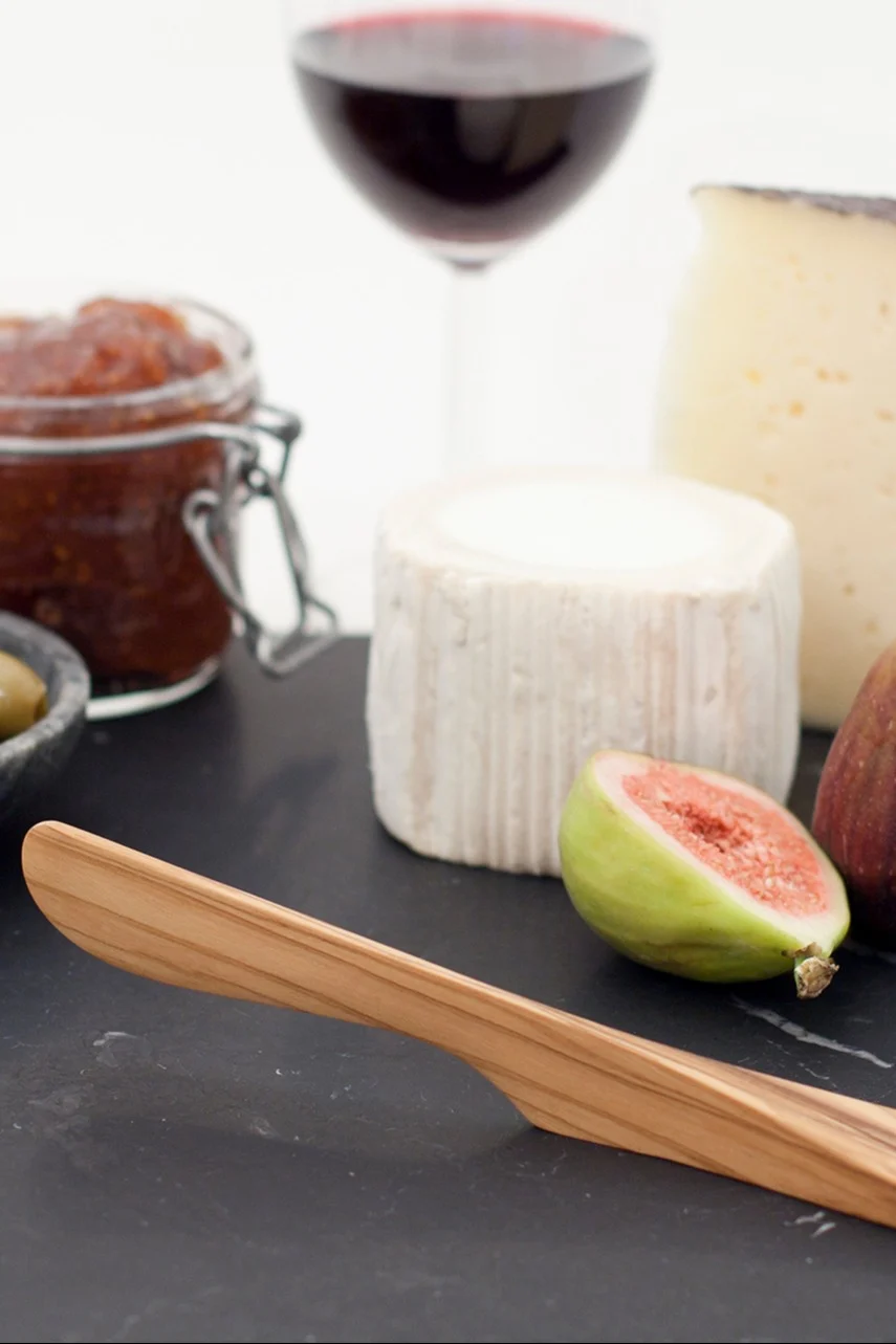 Eine Käseplatte mit einem Holzmesser, zwei Käsesorten, einer halbierten Feige, Marmelade, Oliven und einem Glas Rotwein.