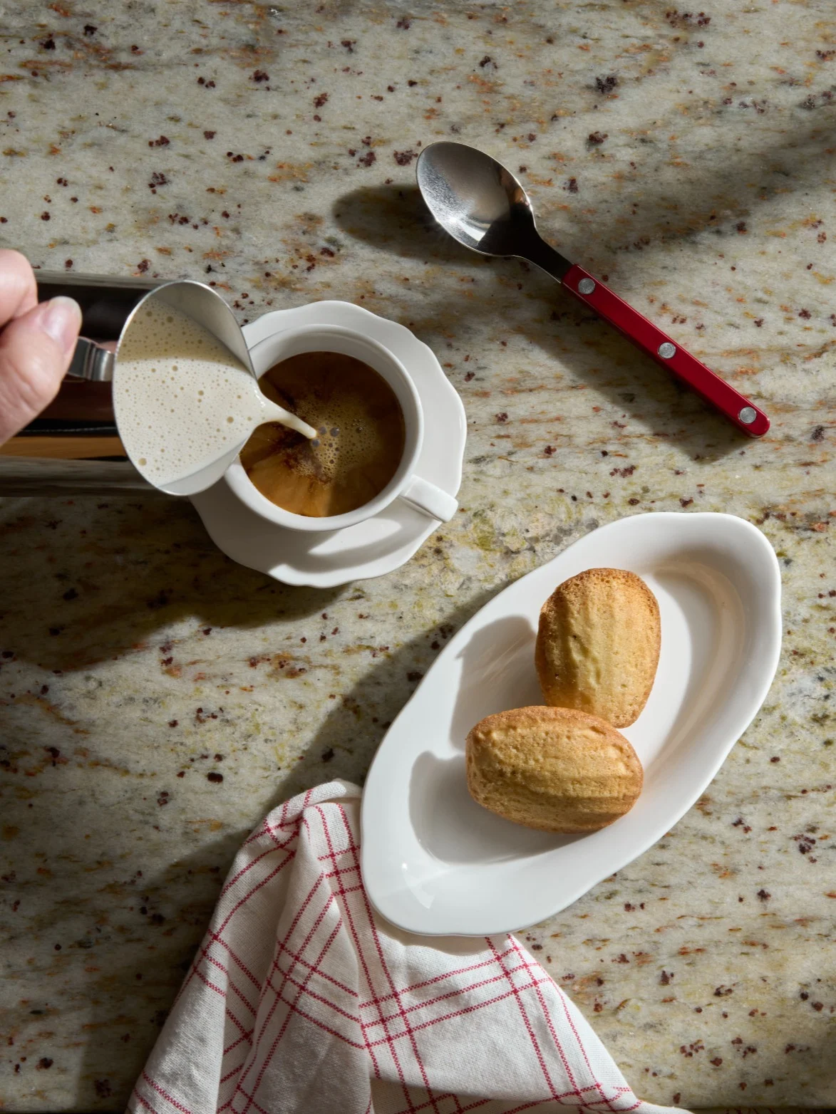 Hand gießt Milchschaum in eine Kaffeetasse neben Madeleines auf einer Granitarbeitsplatte.