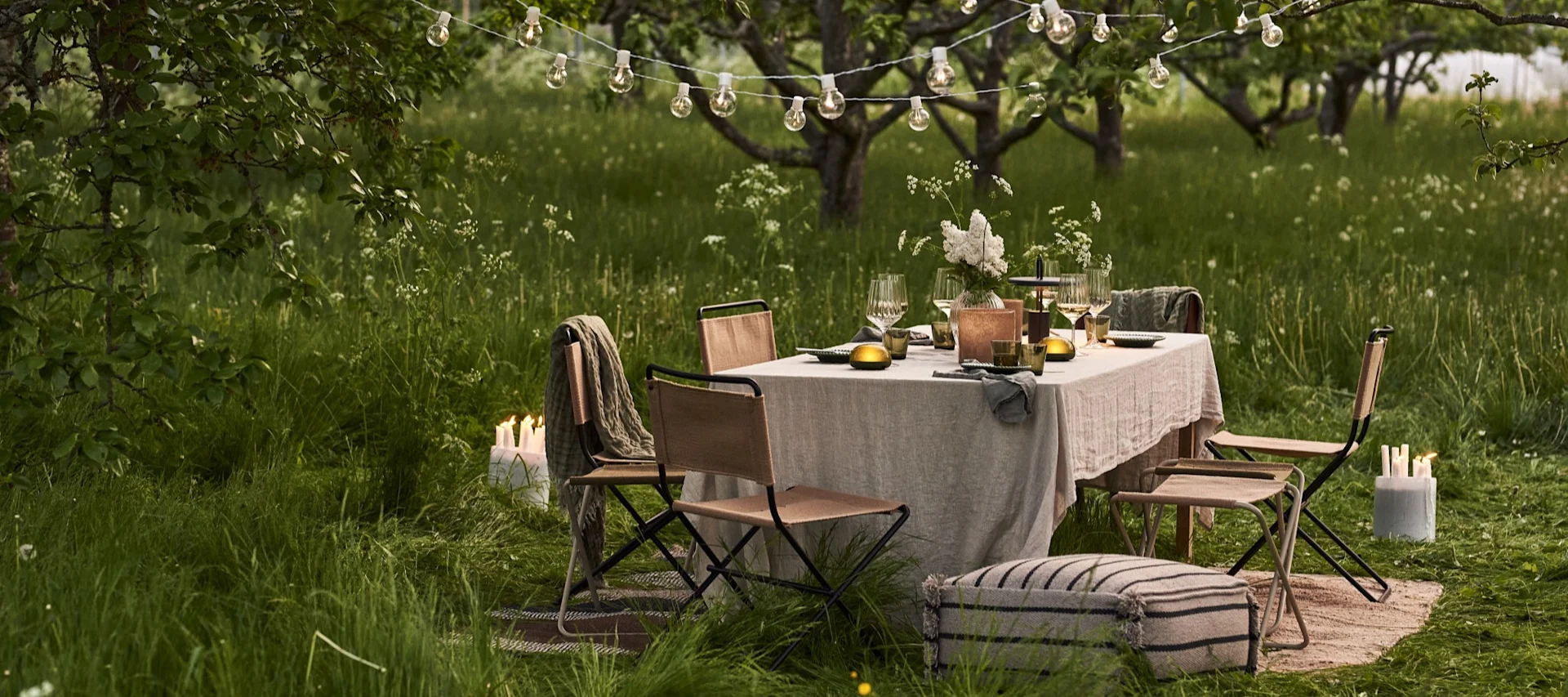 Outdoor-Esstisch unter Lichterketten in einem grasbewachsenen Obstgarten bei Dämmerung, mit Stühlen und Kerzen.