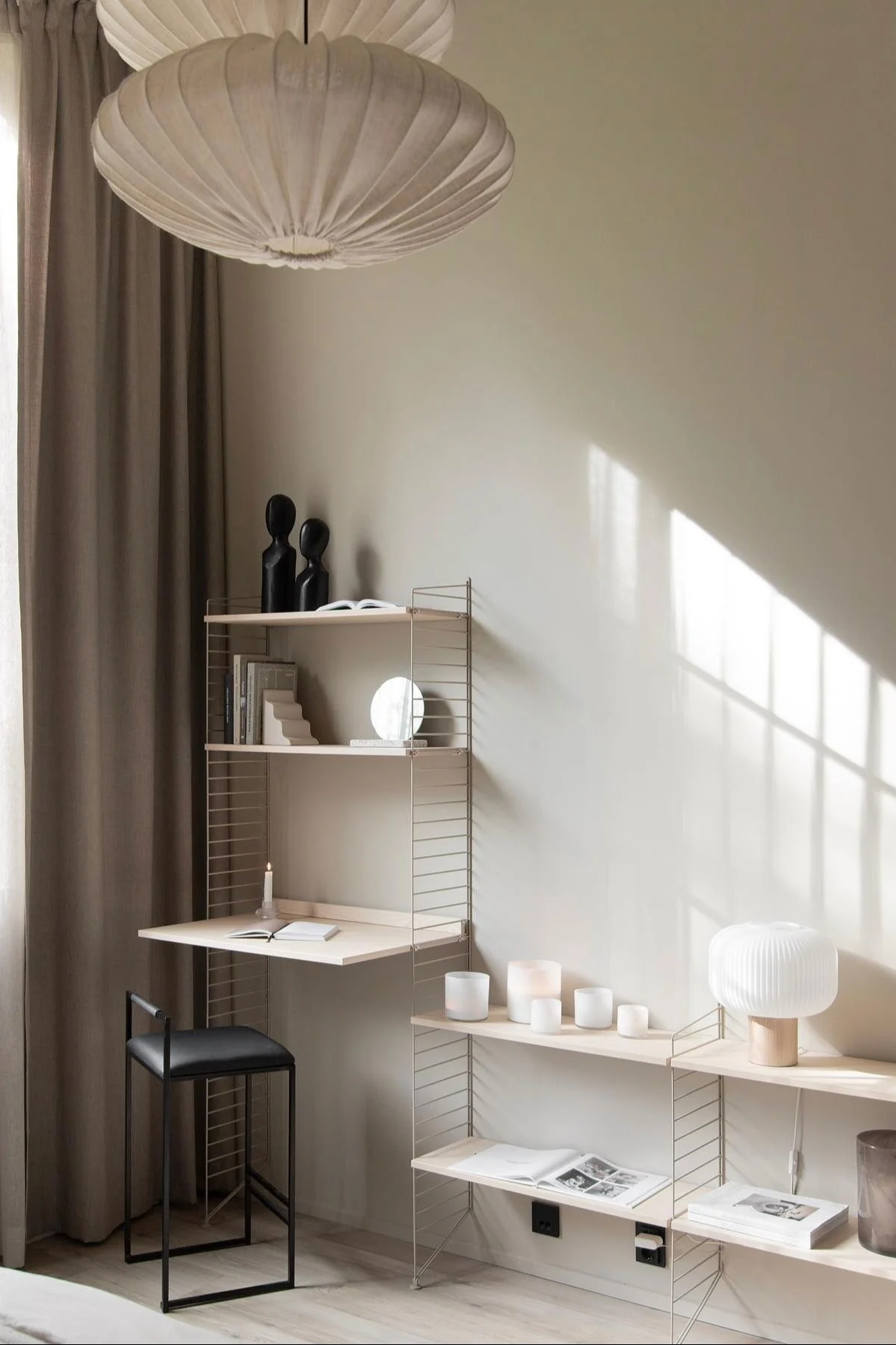 Modernes Homeoffice: Regalsystem aus hellem Holz mit Schreibtisch, schwarzem Hocker und Deko. Sonnenlicht wirft Schatten.