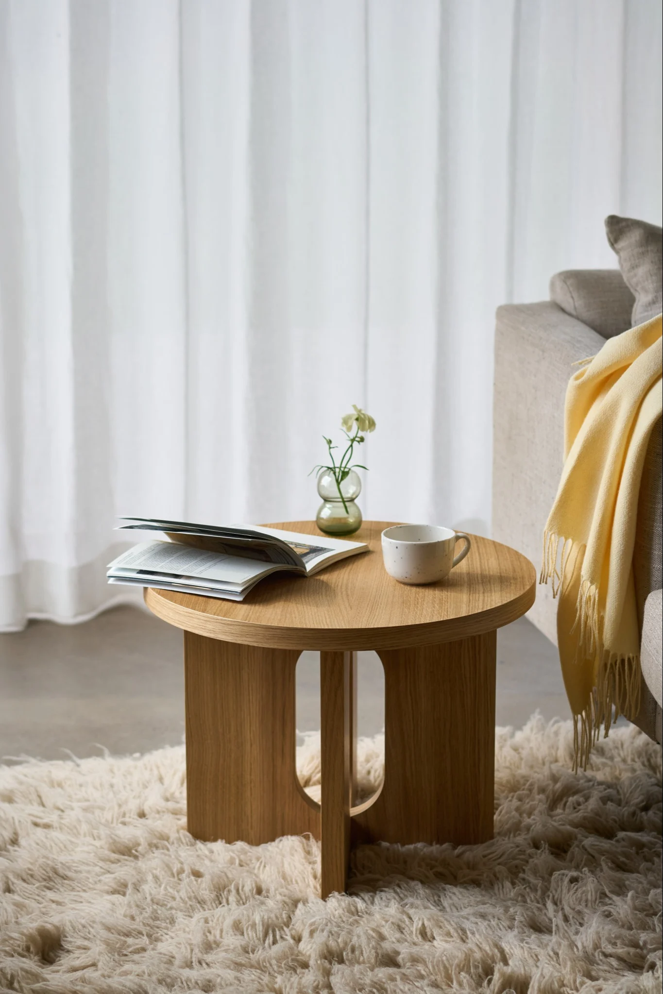 Runder Holztisch mit einem offenen Buch, einer Tasse und einer kleinen Vase mit einer weißen Blume auf einem flauschigen Teppich, neben einem Sofa.