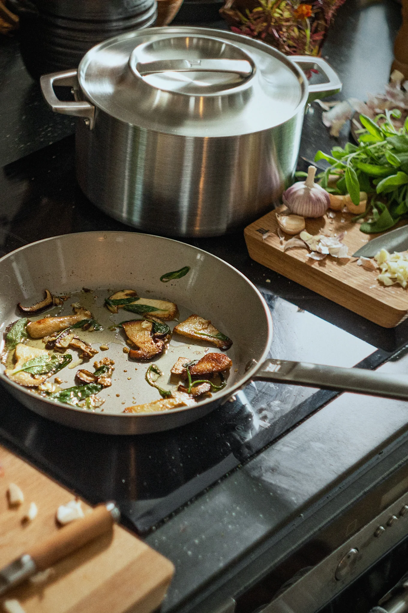 Eine Bratpfanne mit sautierten Pilzen und Salbei auf einem Herd neben einem großen Topf und einem Schneidebrett mit Knoblauch.