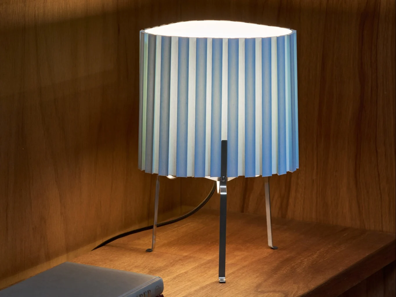 Tischlampe mit plissiertem blau-weiß gestreiftem Schirm auf einem Holzregal, das den Bereich beleuchtet.