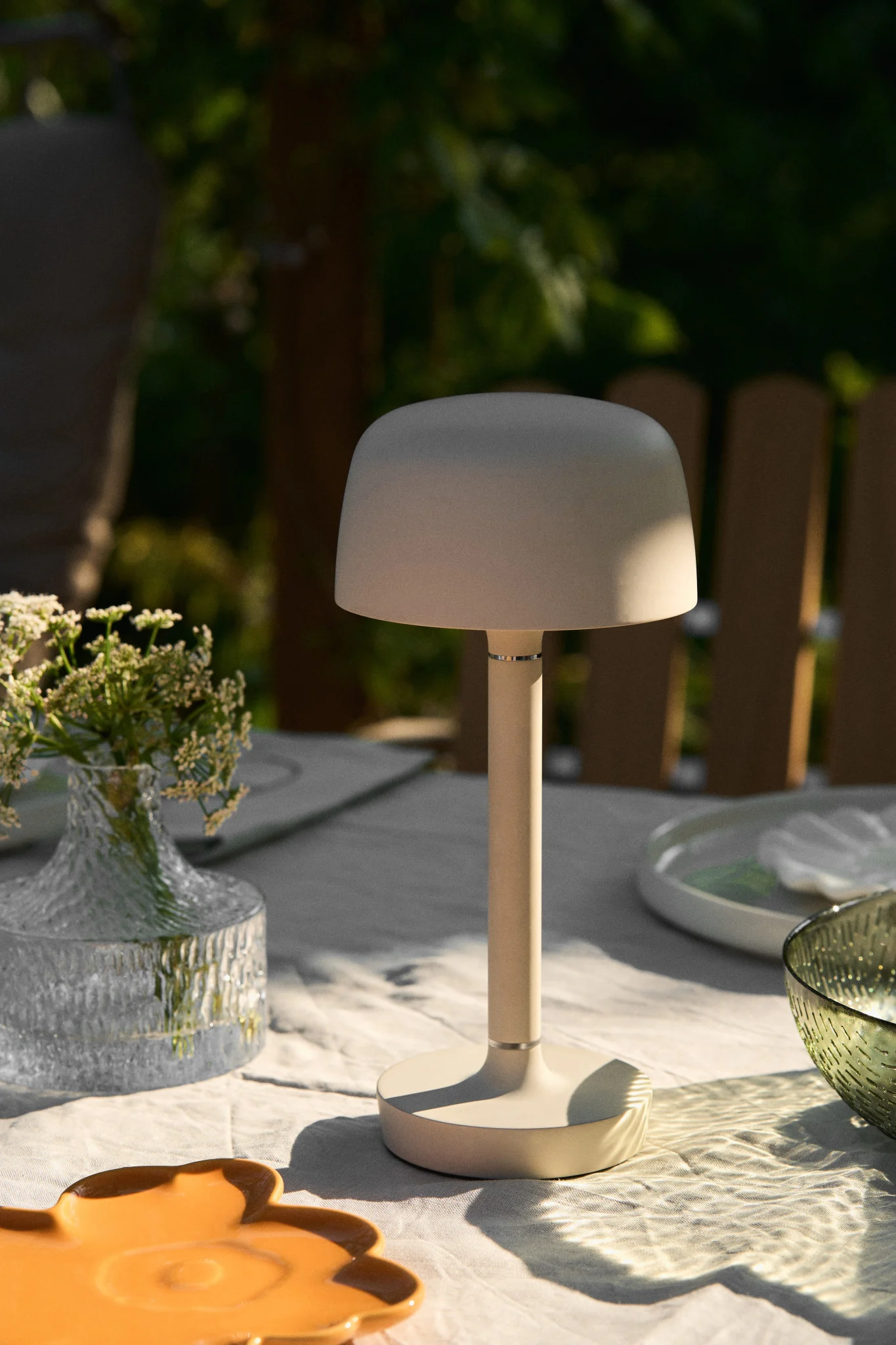 Eine pilzförmige beige Tischlampe auf einem weißen Esstisch im Freien mit Blumen und Geschirr.