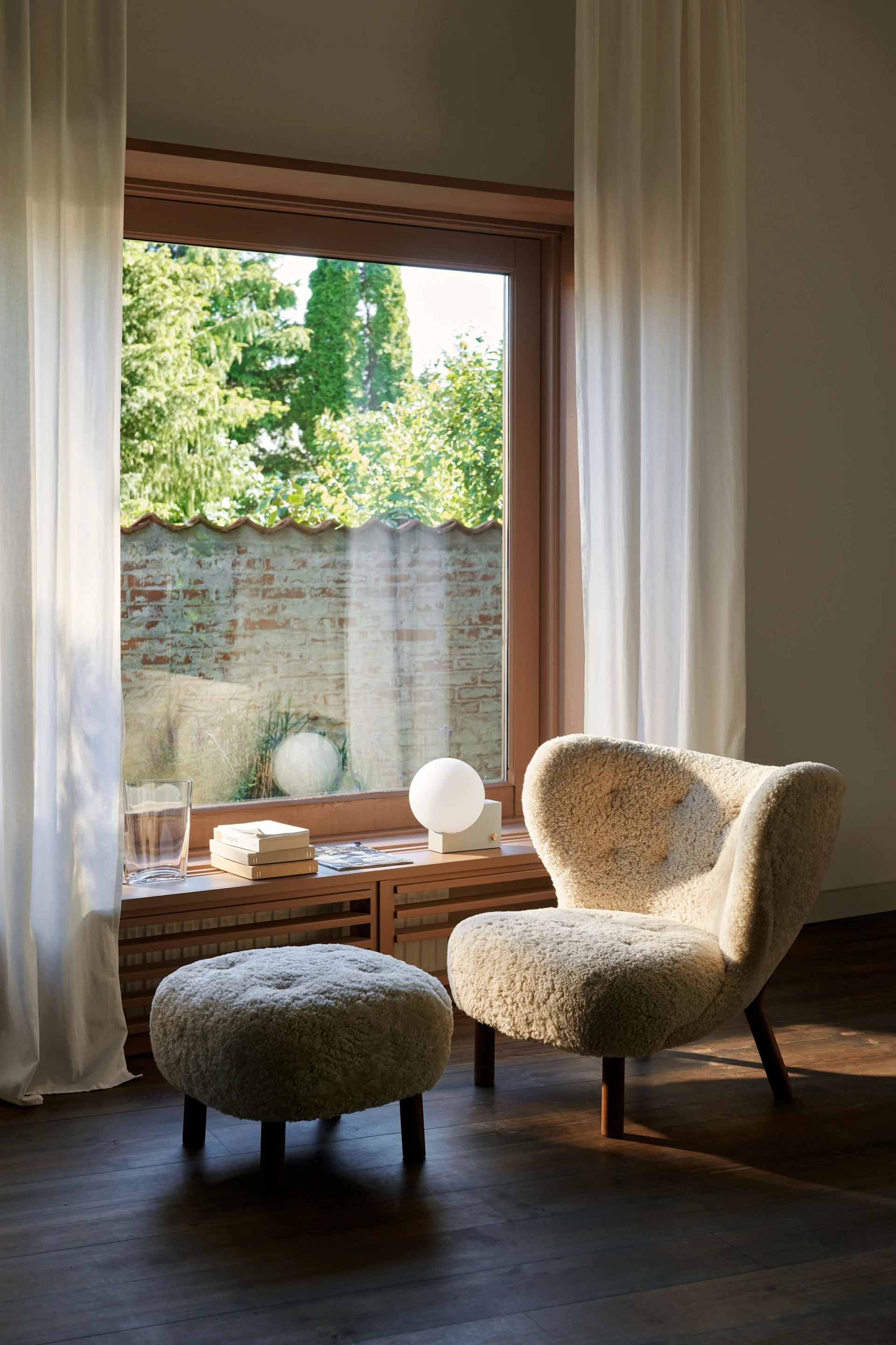 Cremefarbener Bouclé-Sessel und Ottoman am sonnigen Fenster. Auf der Fensterbank: ein Glas, Bücher und eine Kugellampe.