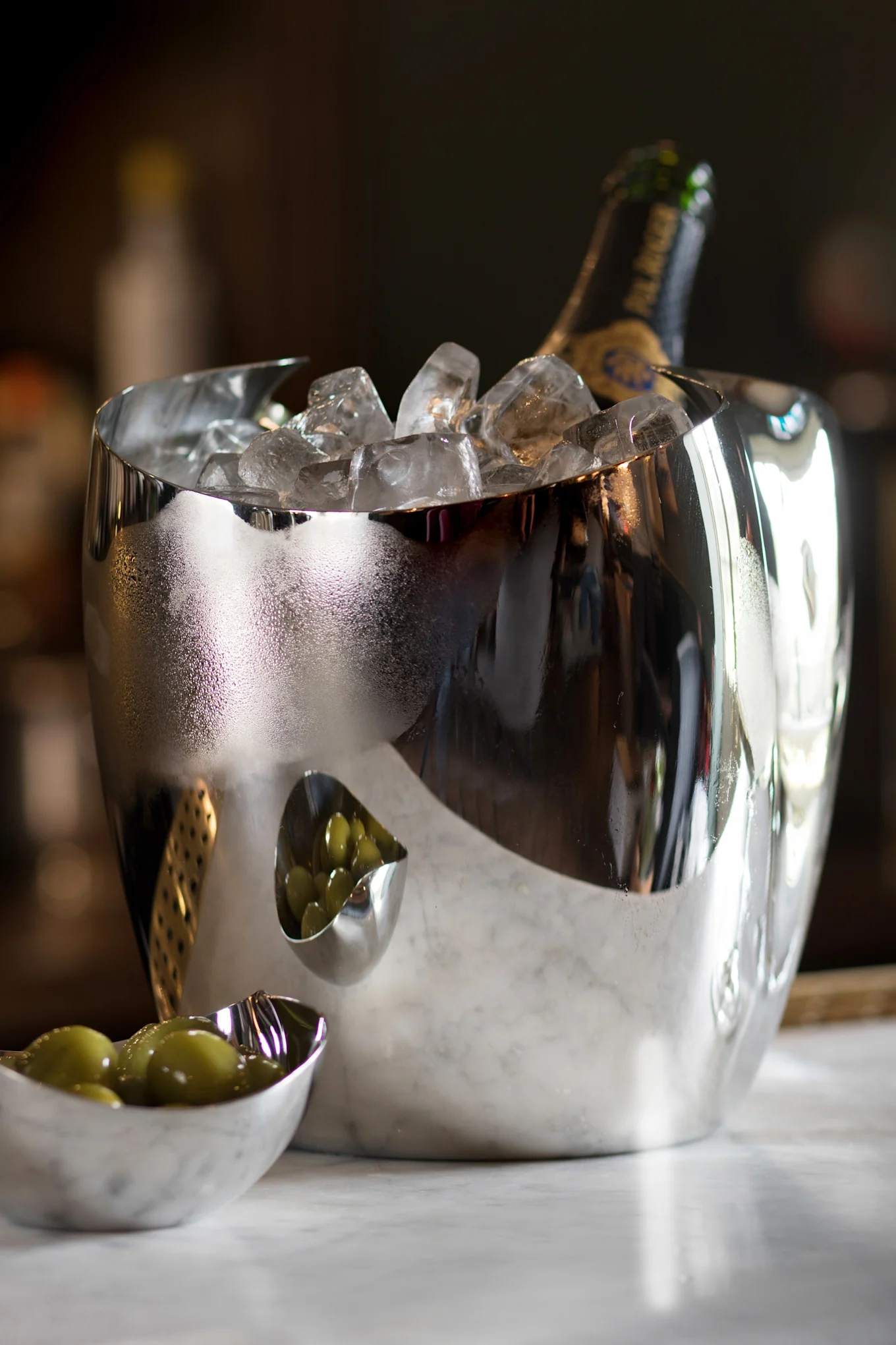 Ein reflektierender silberner Eiskübel mit einer Champagnerflasche und Eis, daneben zwei kleine Schalen mit grünen Oliven auf Marmor.