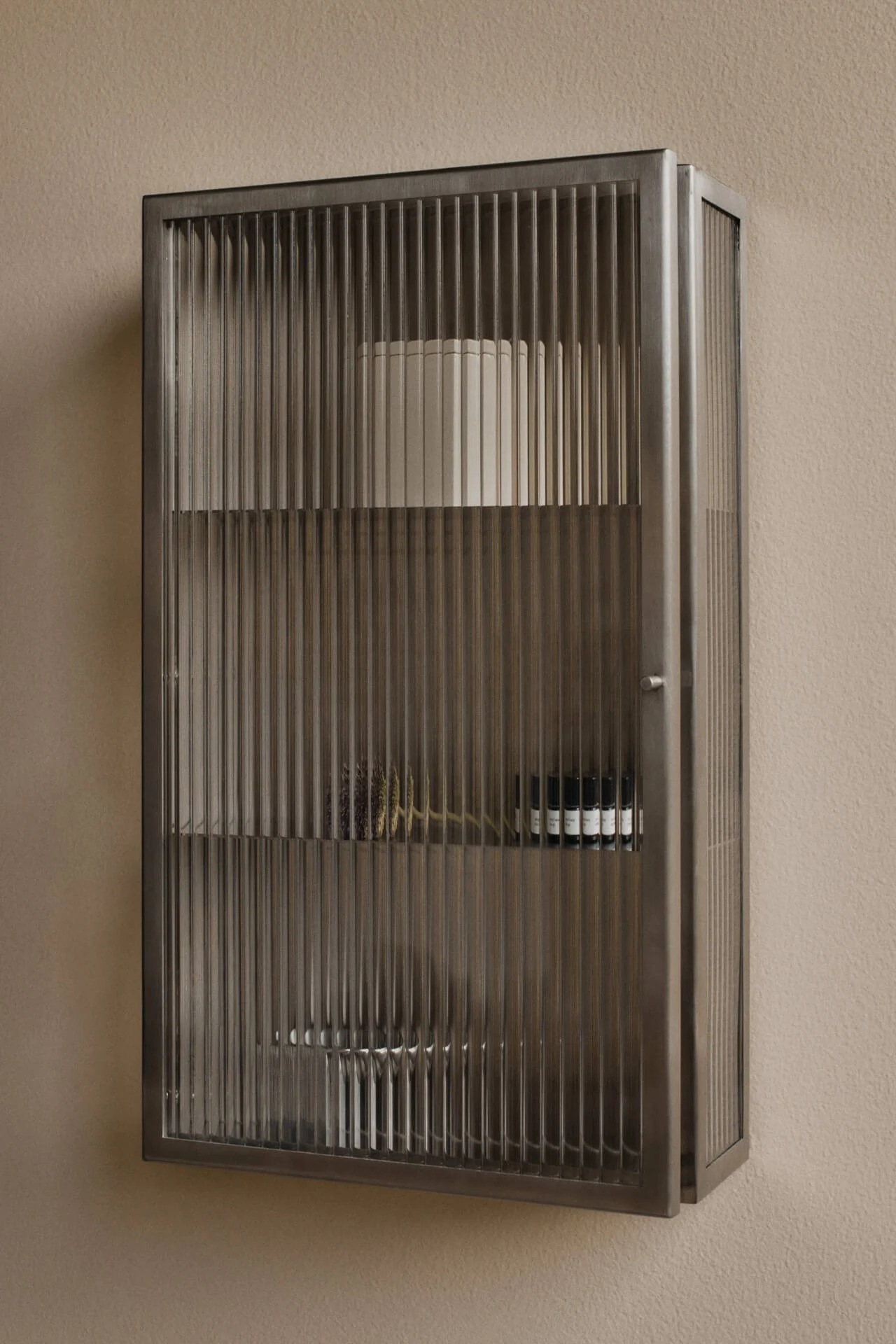 Wandmontierter, dunkler Metallschrank mit geriffelten Glastüren an einer beigefarbenen Wand. Enthält Regale mit einem weißen Zylinder, getrockneten Pflanzen und kleinen dunklen Flaschen.
