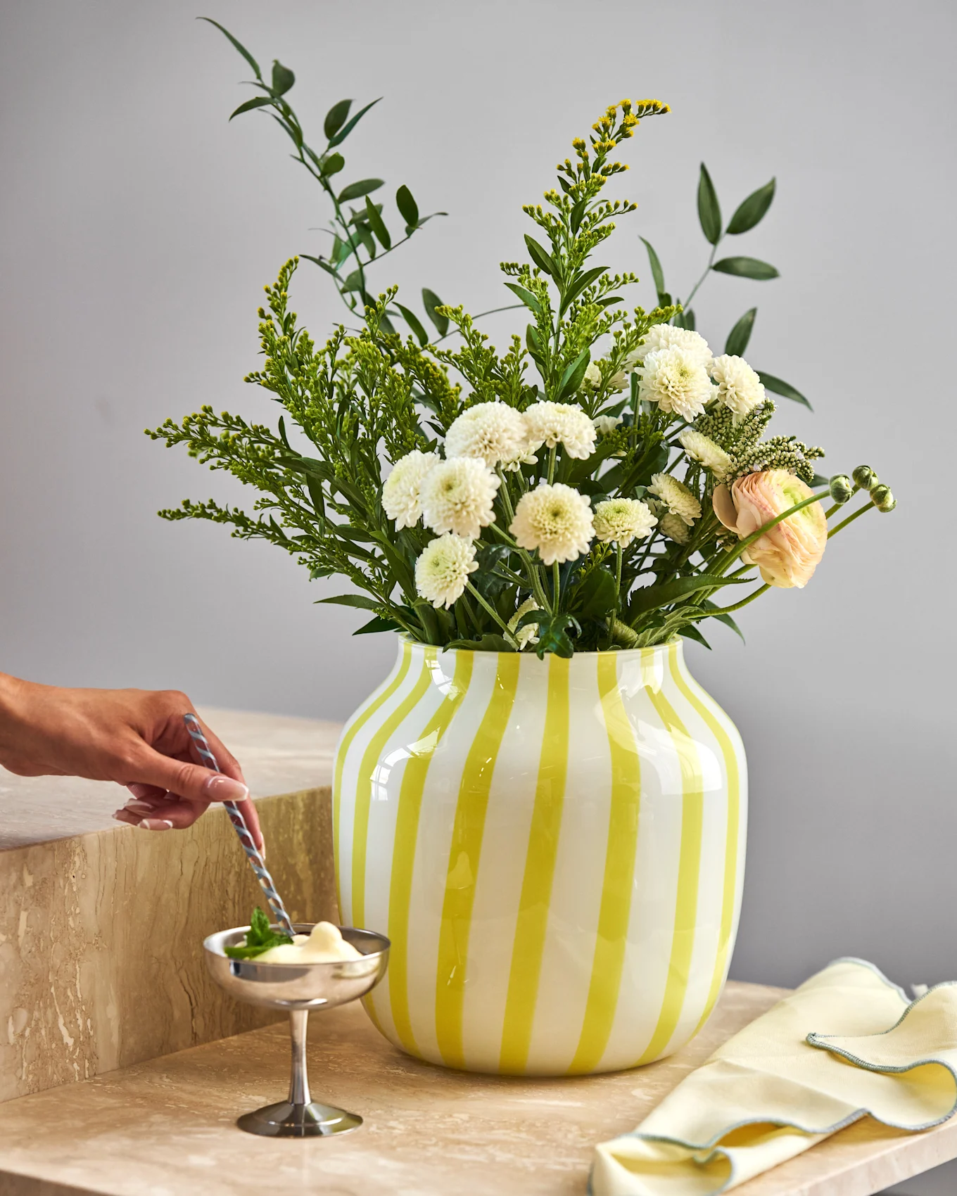 Hand rührt Dessert in Silberschale auf Marmor. Gelb gestreifte Vase mit weißen Chrysanthemen, gelben Blumen und rosa Ranunkeln.