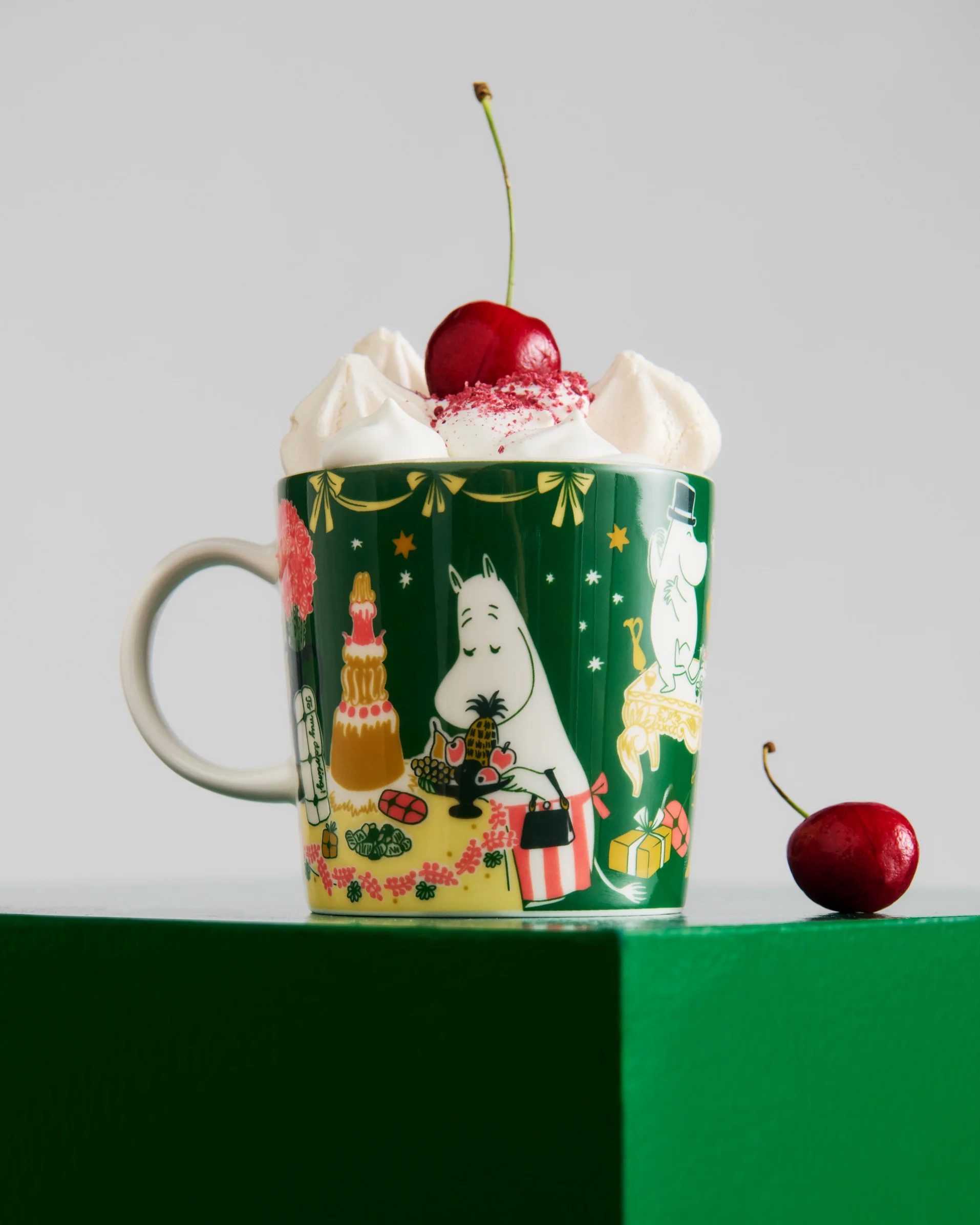 Dunkelgrüne Tasse mit Moomin-Figuren, gekrönt mit Schlagsahne, roten Streuseln und einer Kirsche. Eine weitere Kirsche liegt auf der grünen Oberfläche.