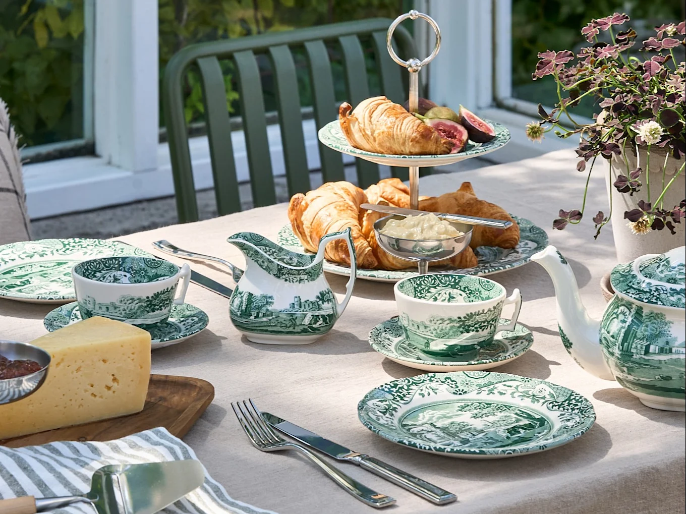Ein Gartentisch mit grün-weißem Geschirr, Croissants, Käse und einer Teekanne in einer Gartenumgebung.