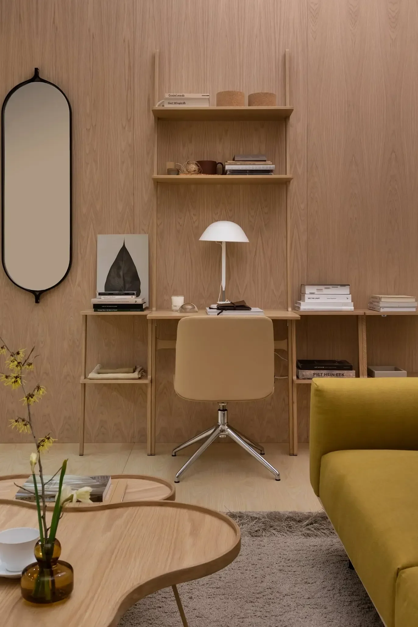 Ein minimalistisches Heimbüro mit heller Holzvertäfelung, beigem Bürostuhl, weißer Lampe, Regalen, neben einem gelben Sofa und Couchtischen.