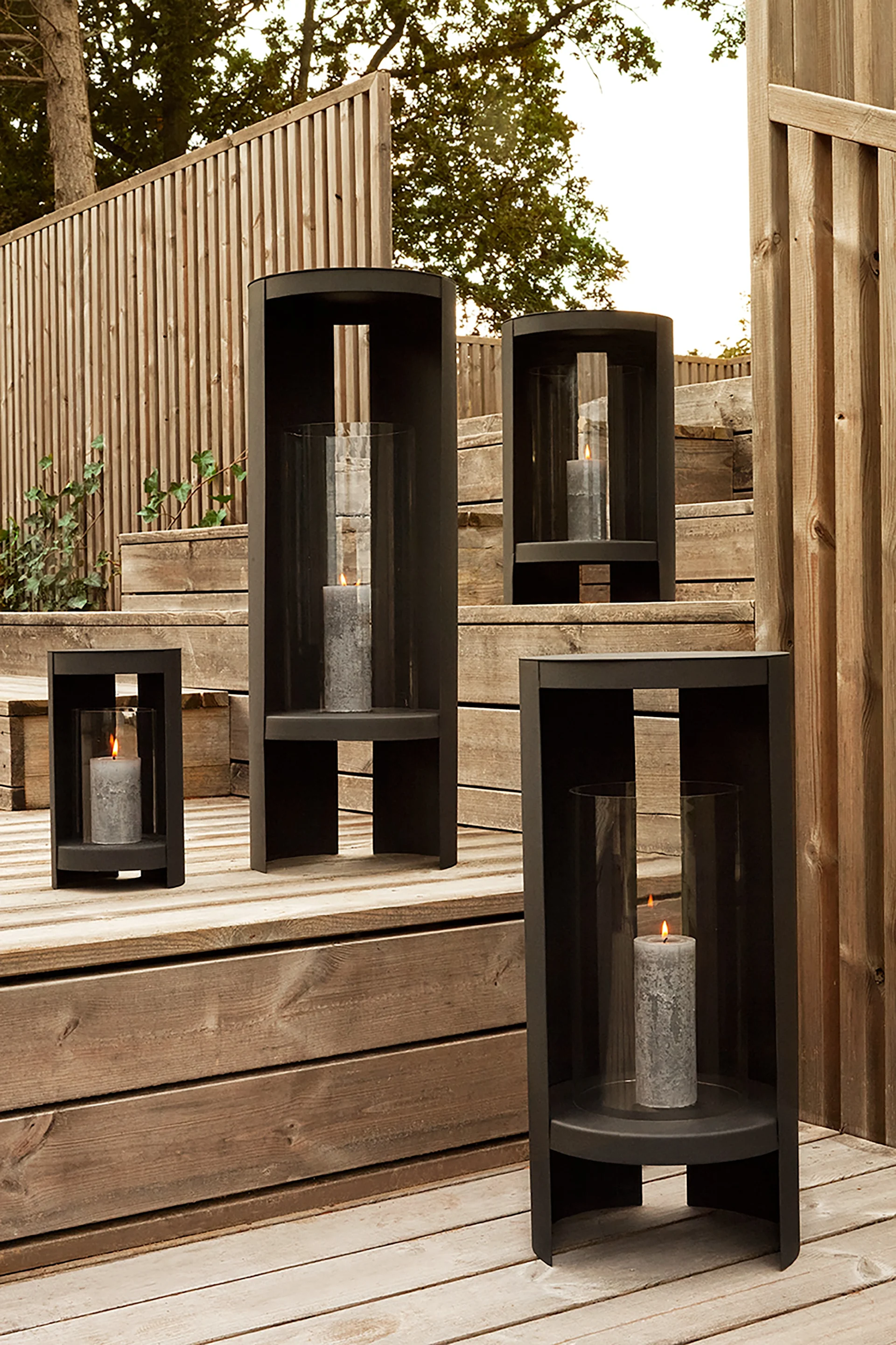 Laternen und Windlichter für draußen – hier sehen Sie die Oro-Laterne von AYTM in Schwarz und in verschiedenen Größen auf einer hölzernen Treppe in einem Garten stehen.
