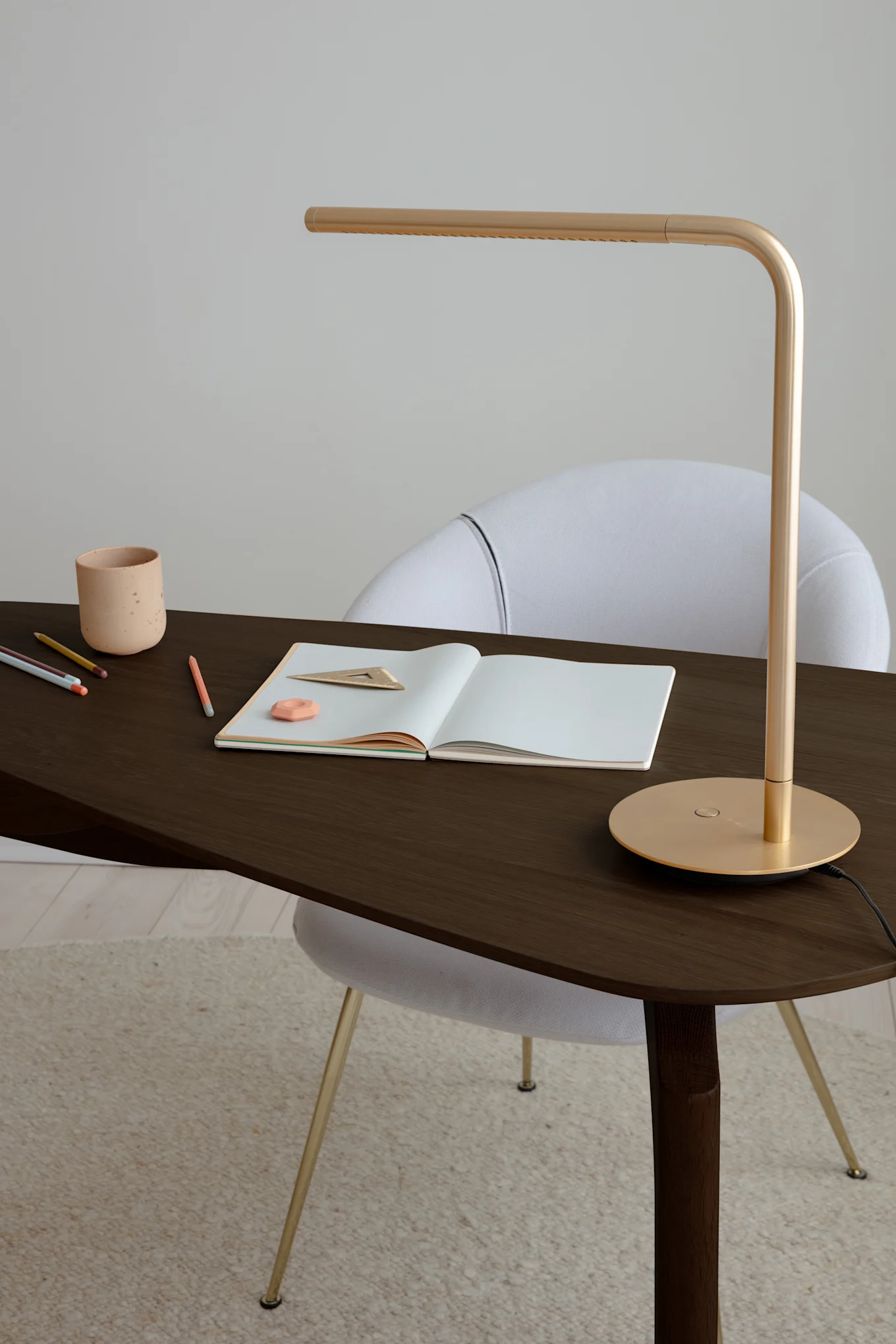 Goldene Schreibtischlampe auf dunklem Holztisch mit Notizbuch, Stiften, Radiergummi, Lineal und Keramiktasse. Weißer Stuhl dahinter.