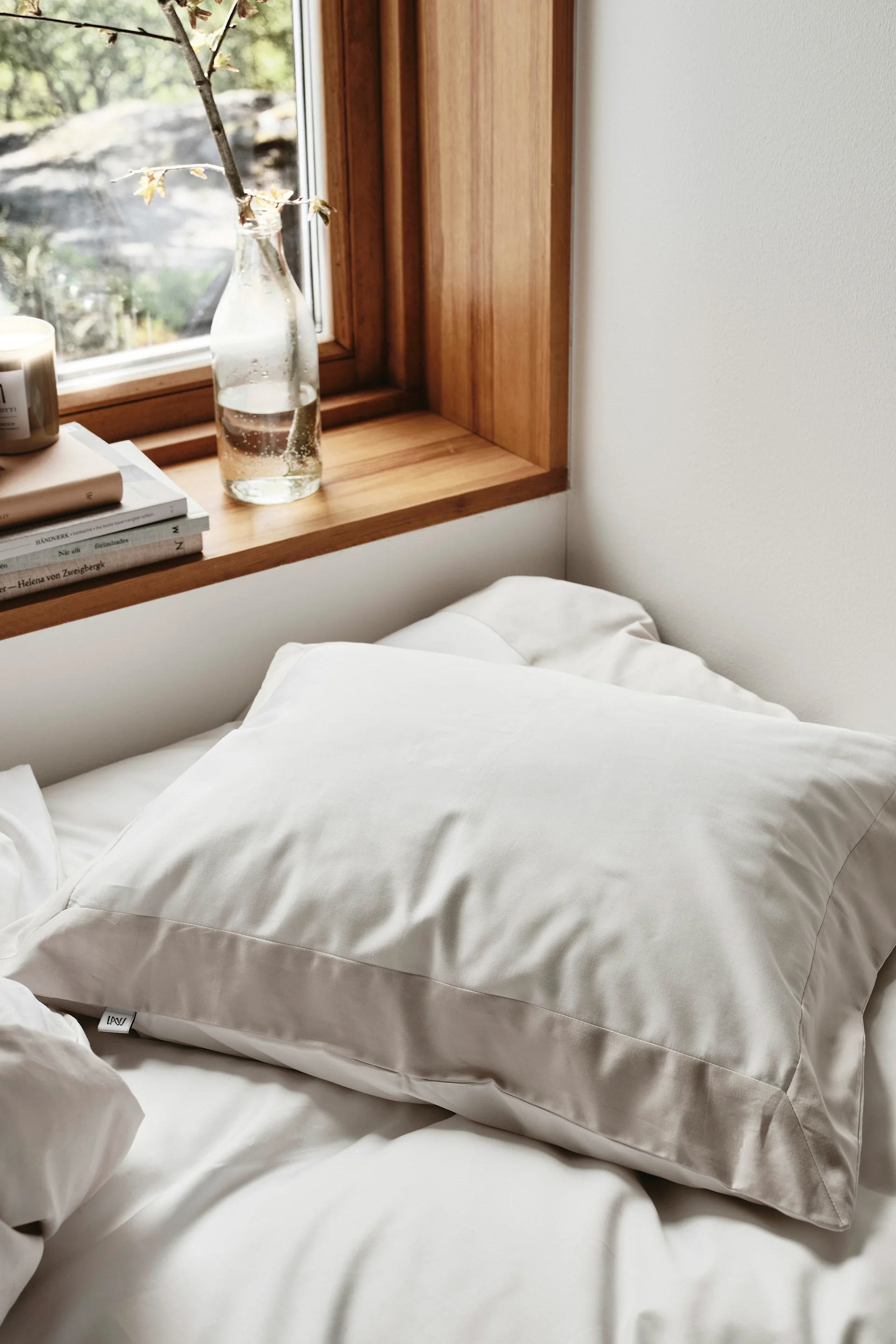 Ein hellbeiges Kissen auf einem weißen Bett, neben einem Fenster mit Holzfensterbank, Büchern und einem Pflanzenzweig in einer Flasche.