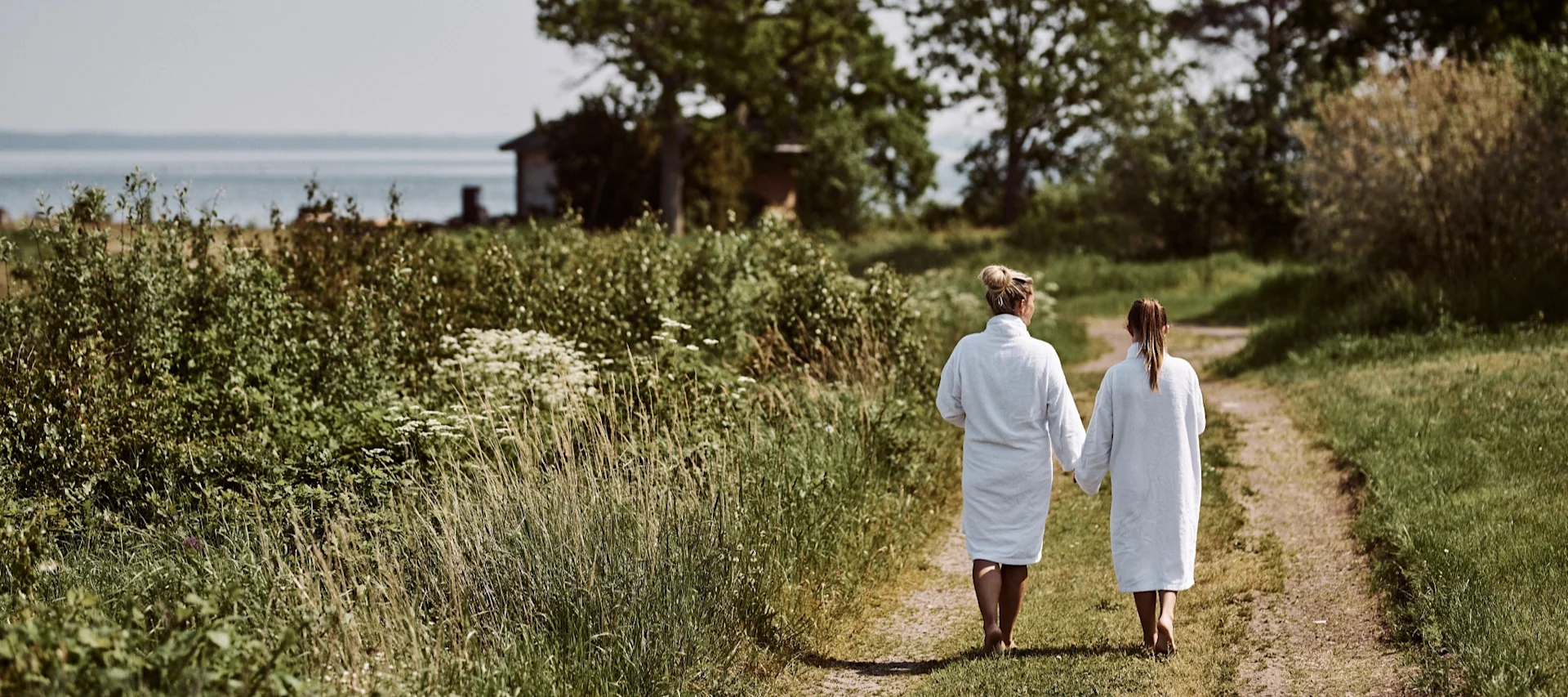 Summer Bucket List: Eine Frau mit einem Mädchen an der Hand tragen weiße Bademäntel und spazieren einen Weg am Meer entlang.