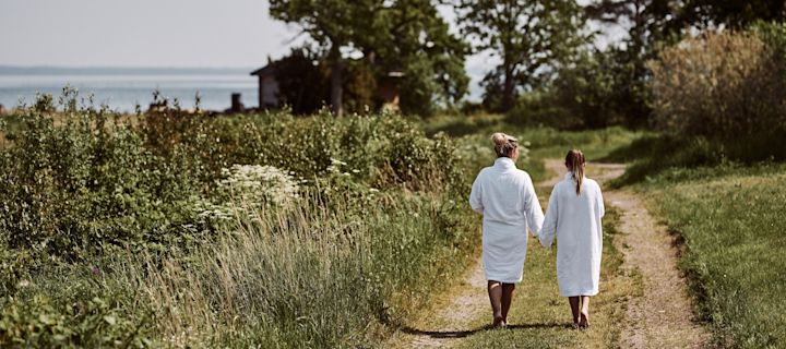 Summer Bucket List: Eine Frau mit einem Mädchen an der Hand tragen weiße Bademäntel und spazieren einen Weg am Meer entlang.