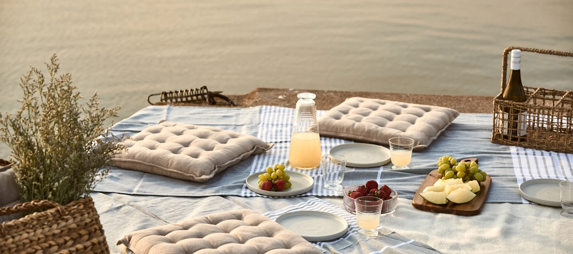 Picknick am Wasser mit Kissen, Obst und Getränken auf einer Decke.