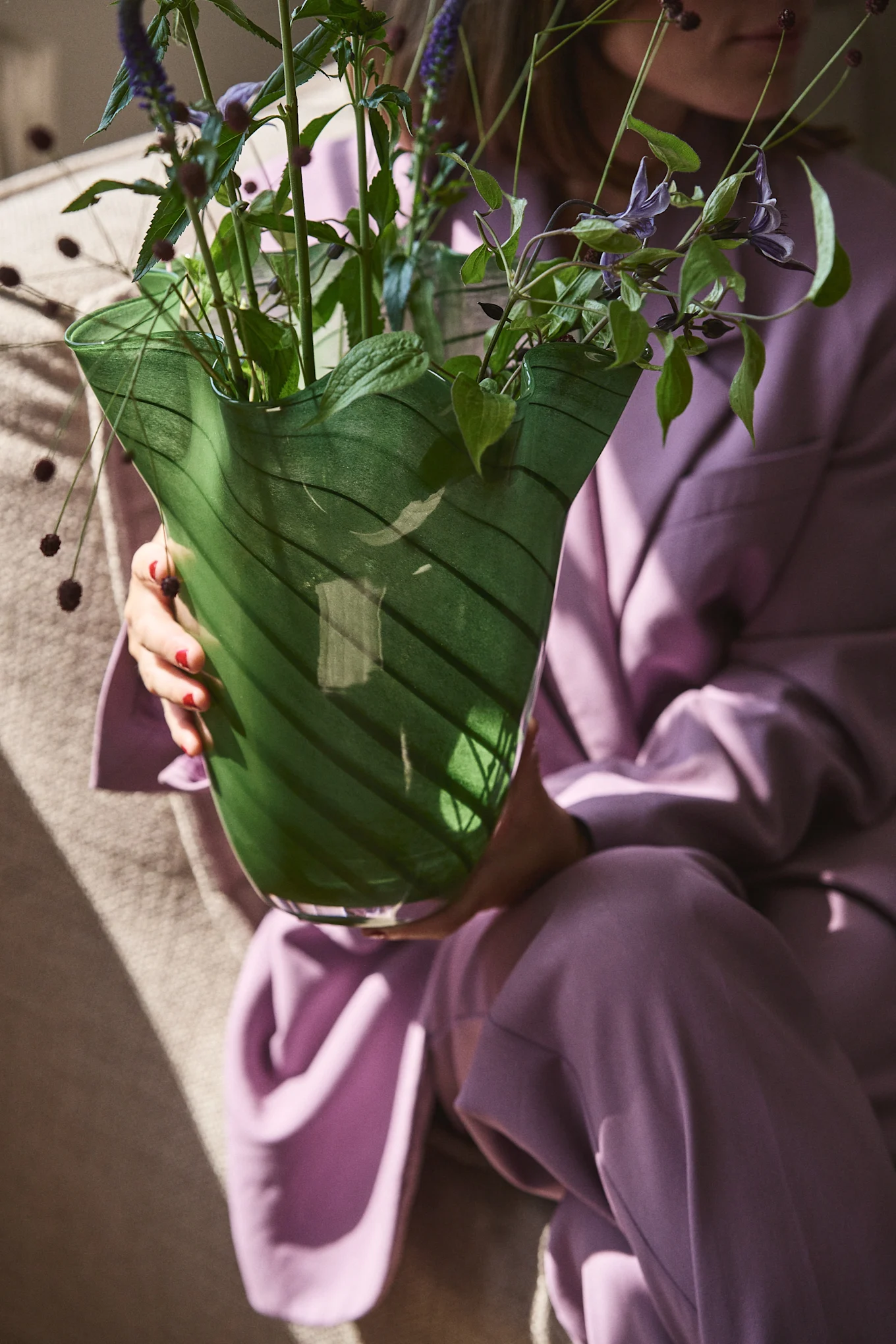Hier sehen Sie eine Person in violetter Kleidung, die die grüne Glasvase Tiggy von Byon hält, eine Vase mit gefalteter, glockenförmiger Form in Erbsengrün mit Streifen.