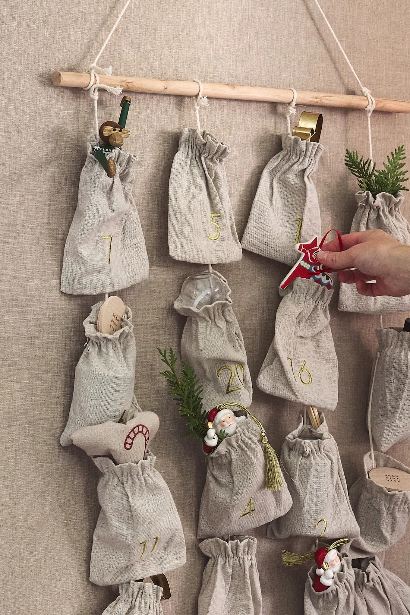 Stoff-Adventskalender mit nummerierten Beuteln an einer Holzstange, gefüllt mit Weihnachtsschmuck; eine Hand legt ein rotes Dalahorse hinein.