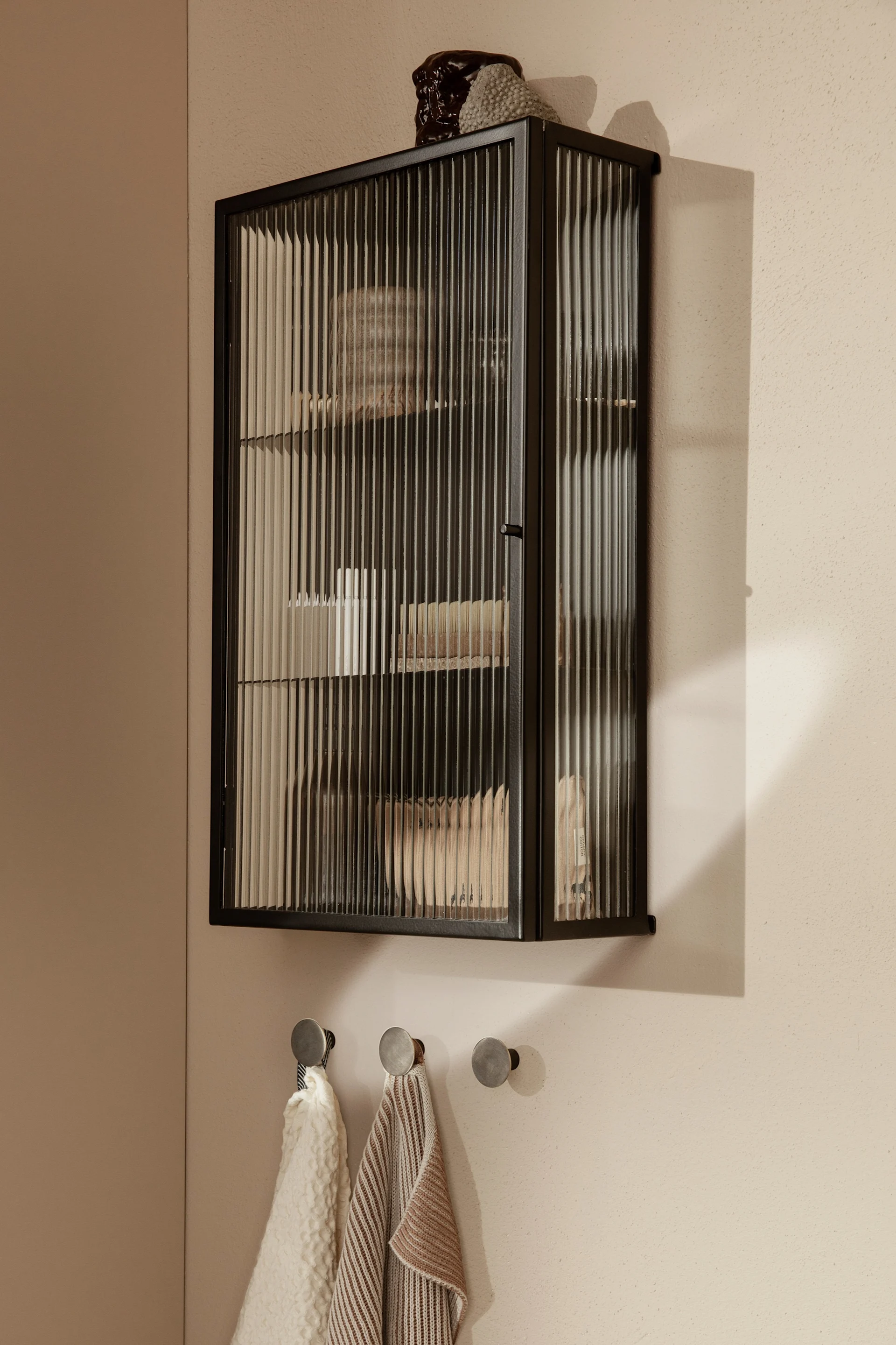 Dunkler Wandschrank mit geriffelten Glastüren, Inhalt zeigend, über drei Metallhaken mit zwei gefalteten Handtüchern.