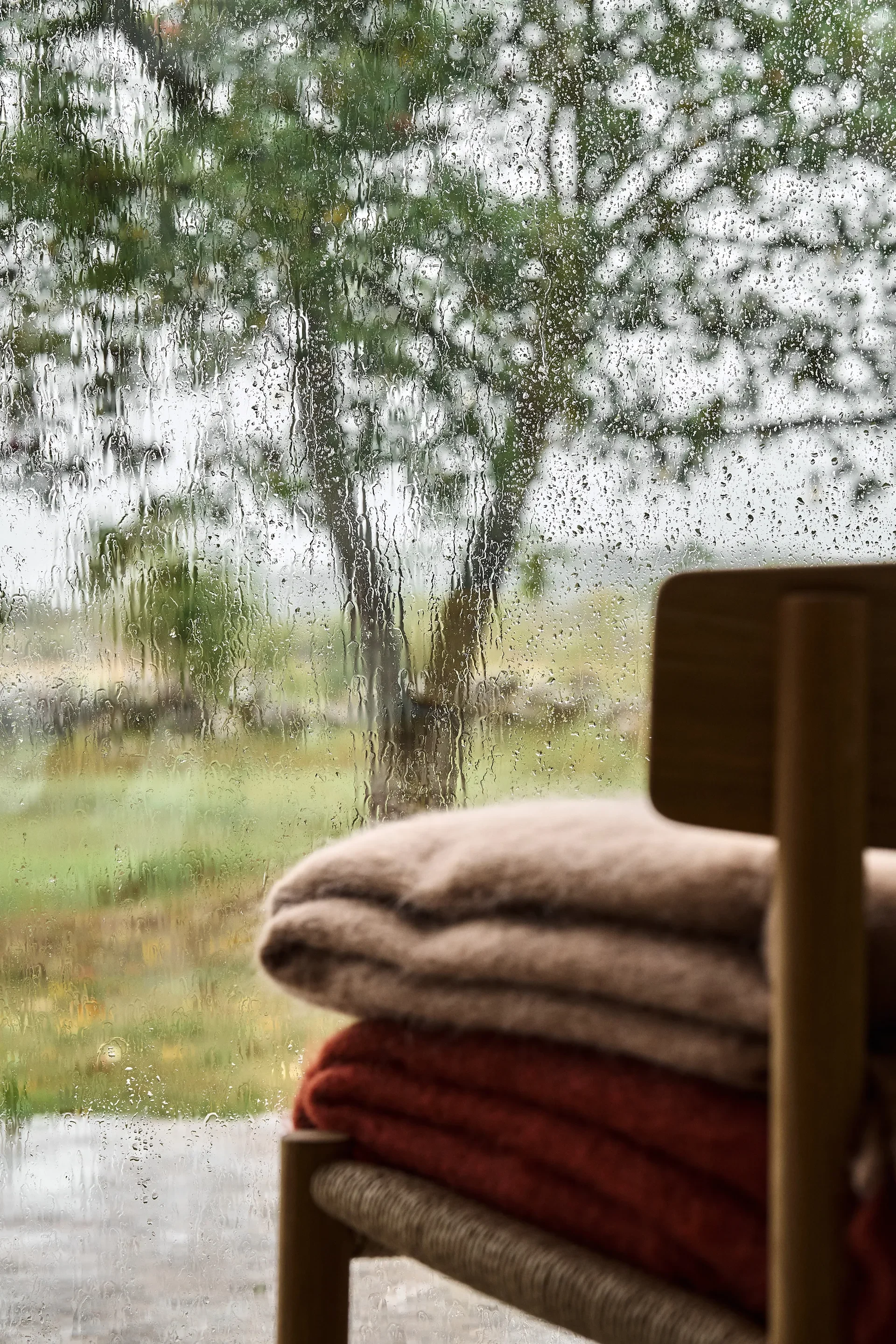 Gestapelte Decken auf einem Holzstuhl an einem regennassen Fenster mit verschwommenen Bäumen.